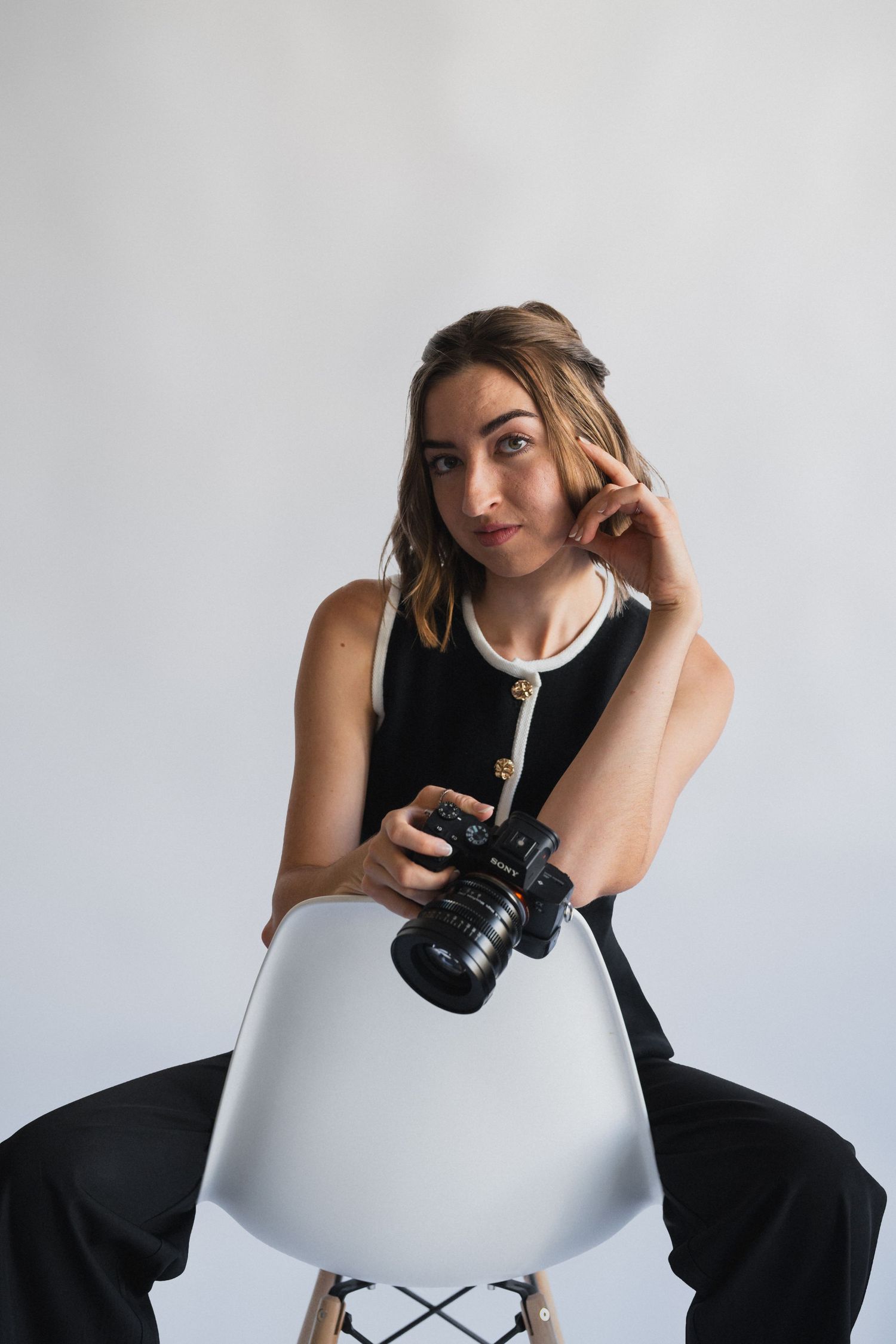 Photographer and filmmaker sits casually on modern white chair while holding professional camera.