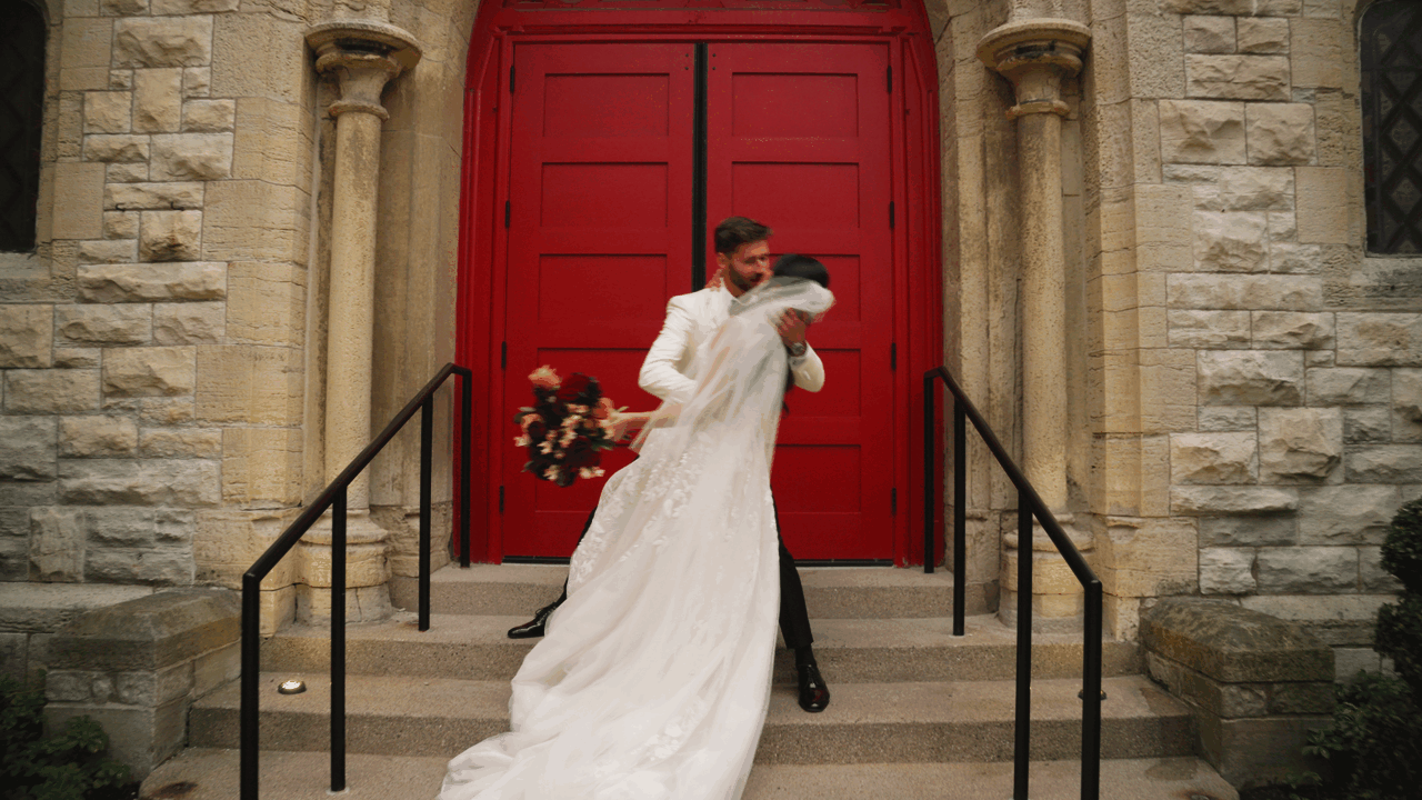 Wedding film of a newly married couple dip kissing in front of red church doors.