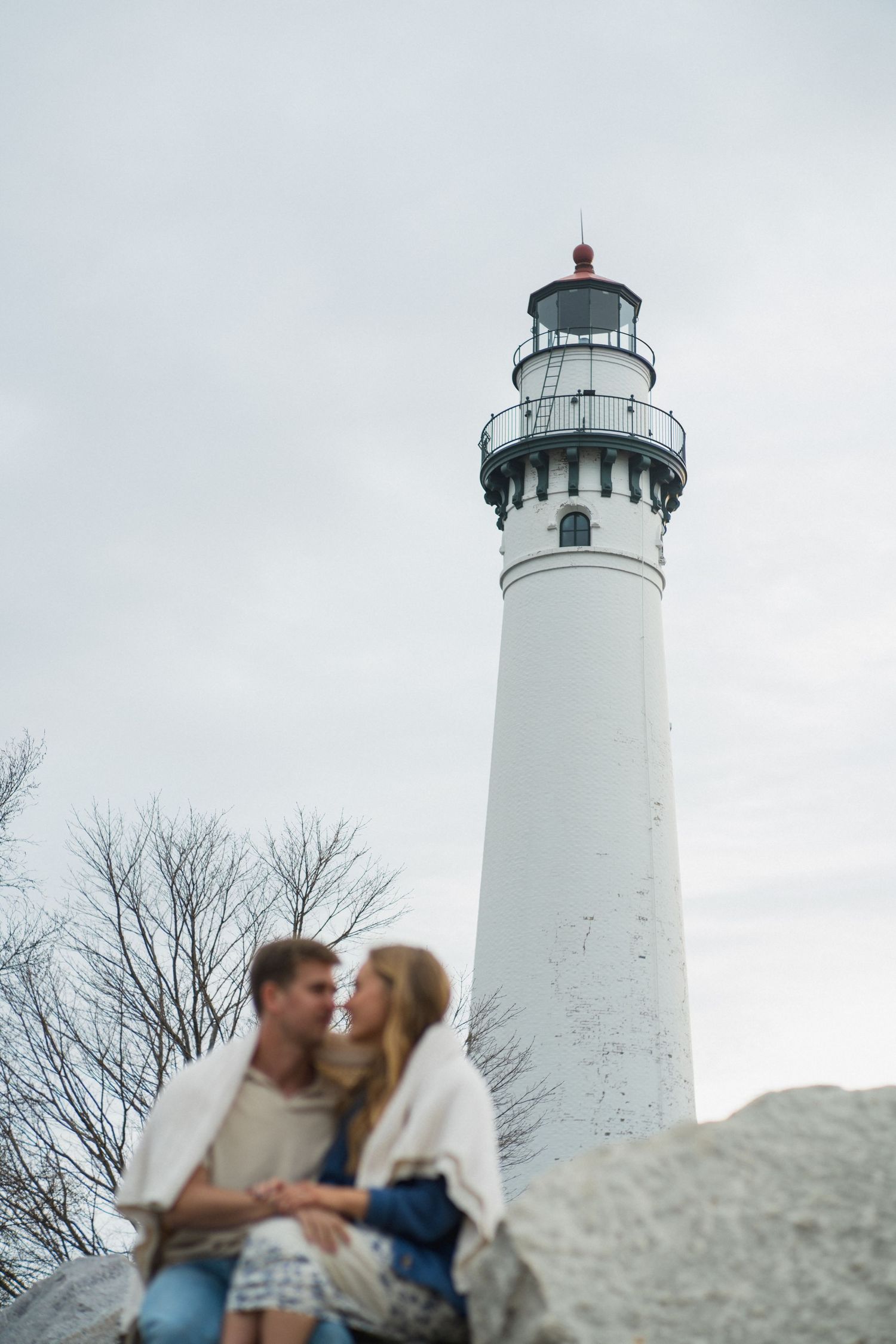 Romantic couple wrapped in blanket sharing a kiss in front of a tall white lighthouse on an overcast day.