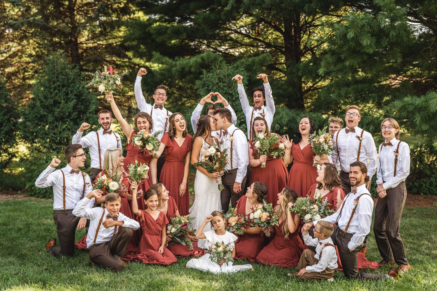 Wedding party makes fun poses and gestures while gathered on grass for group photo.
