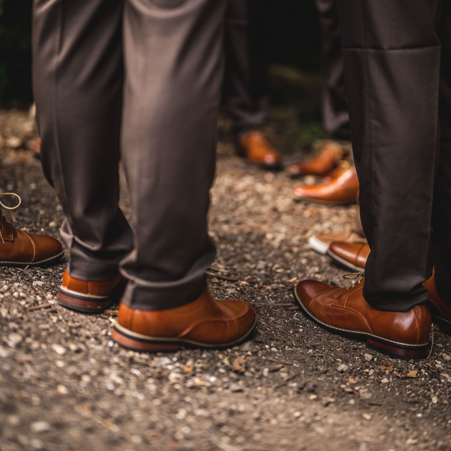 Close-up of brown leather dress shoes worn with dark pants on gravel ground.