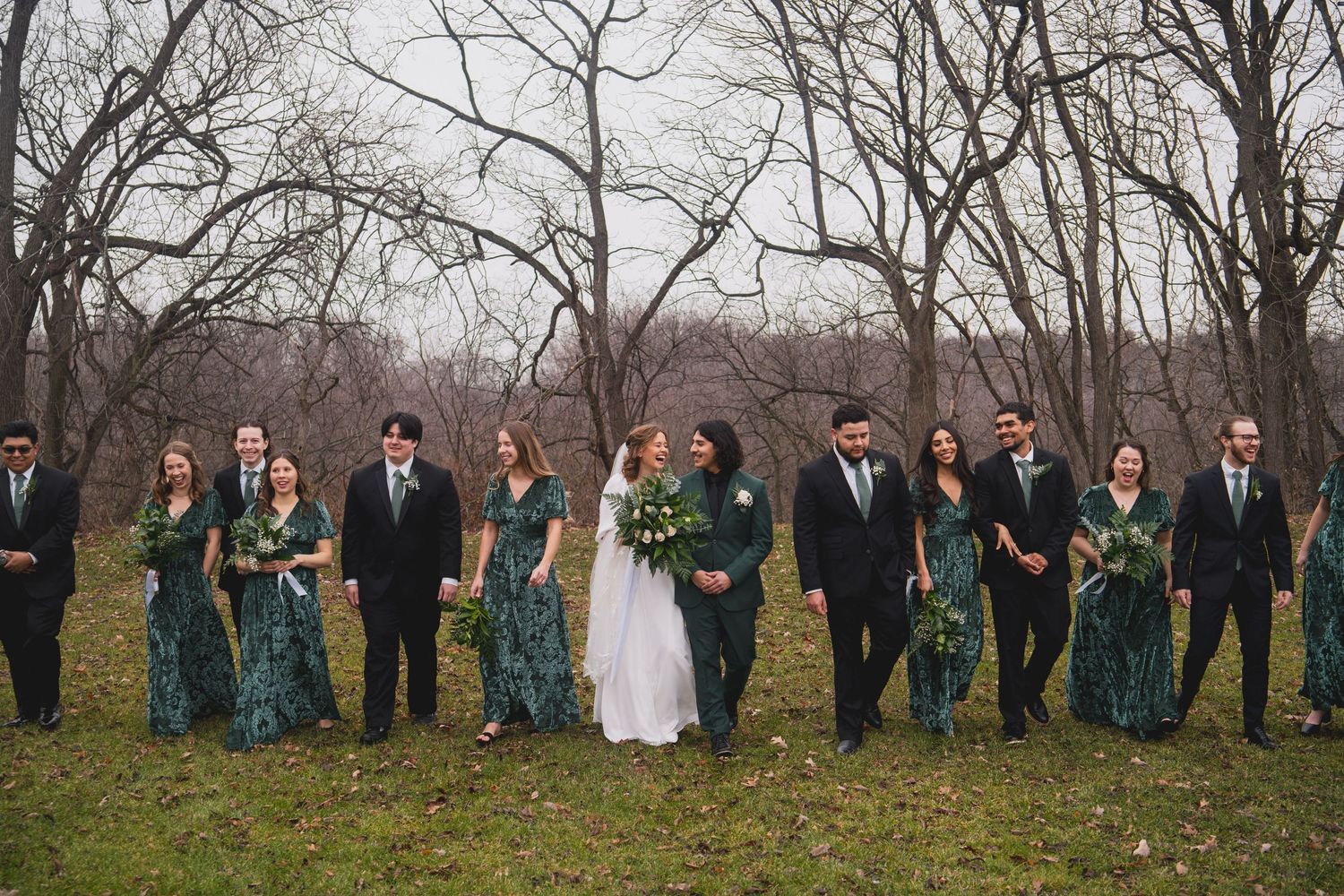 Wedding party in emerald green dresses and black suits poses together in autumn woodland setting.