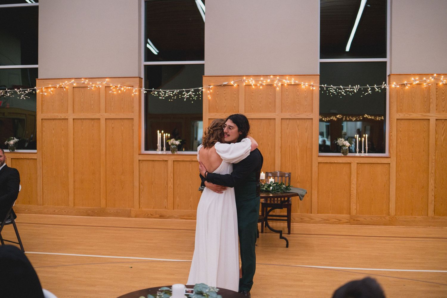 A couple embraces during their wedding dance in a hall decorated with string lights.