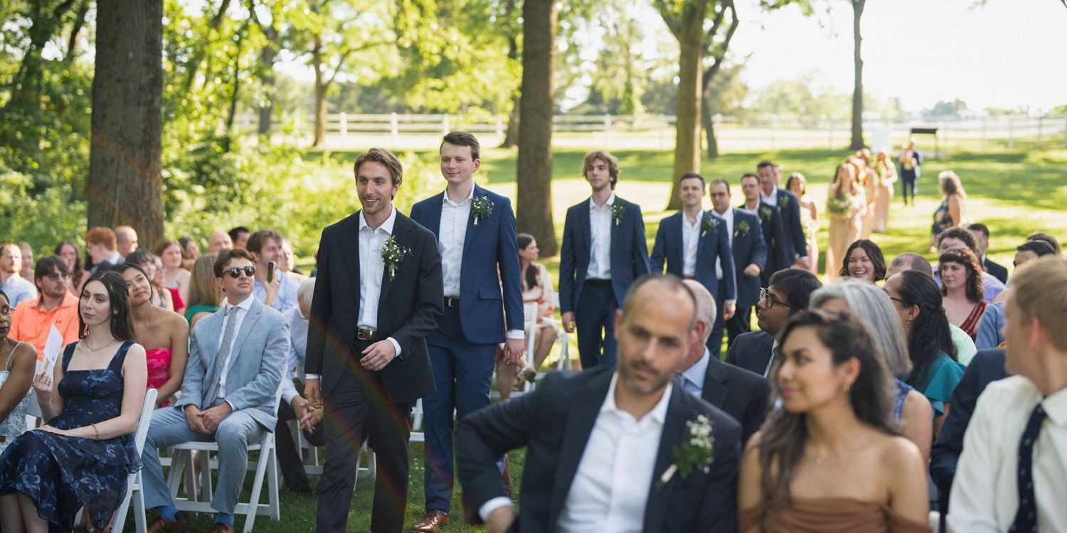 Groomsmen walking down the aisle at an outdoor wedding ceremony with guests seated on white chairs.