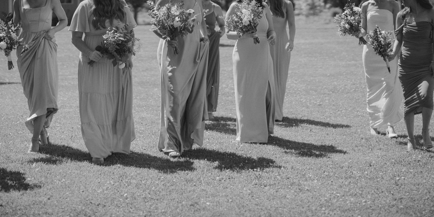 Black and white photo of bridesmaids in long gowns holding bouquets walking across a grassy outdoor wedding venue.