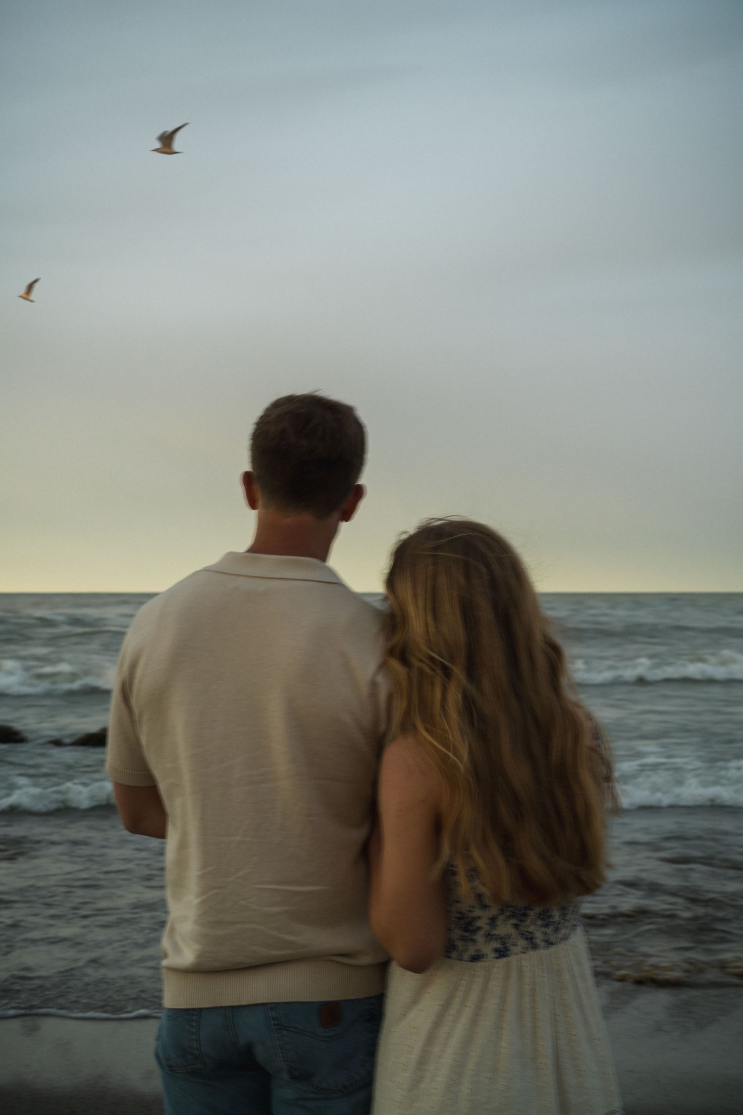 Couple standing together watching ocean waves at dusk with seagulls flying overhead.