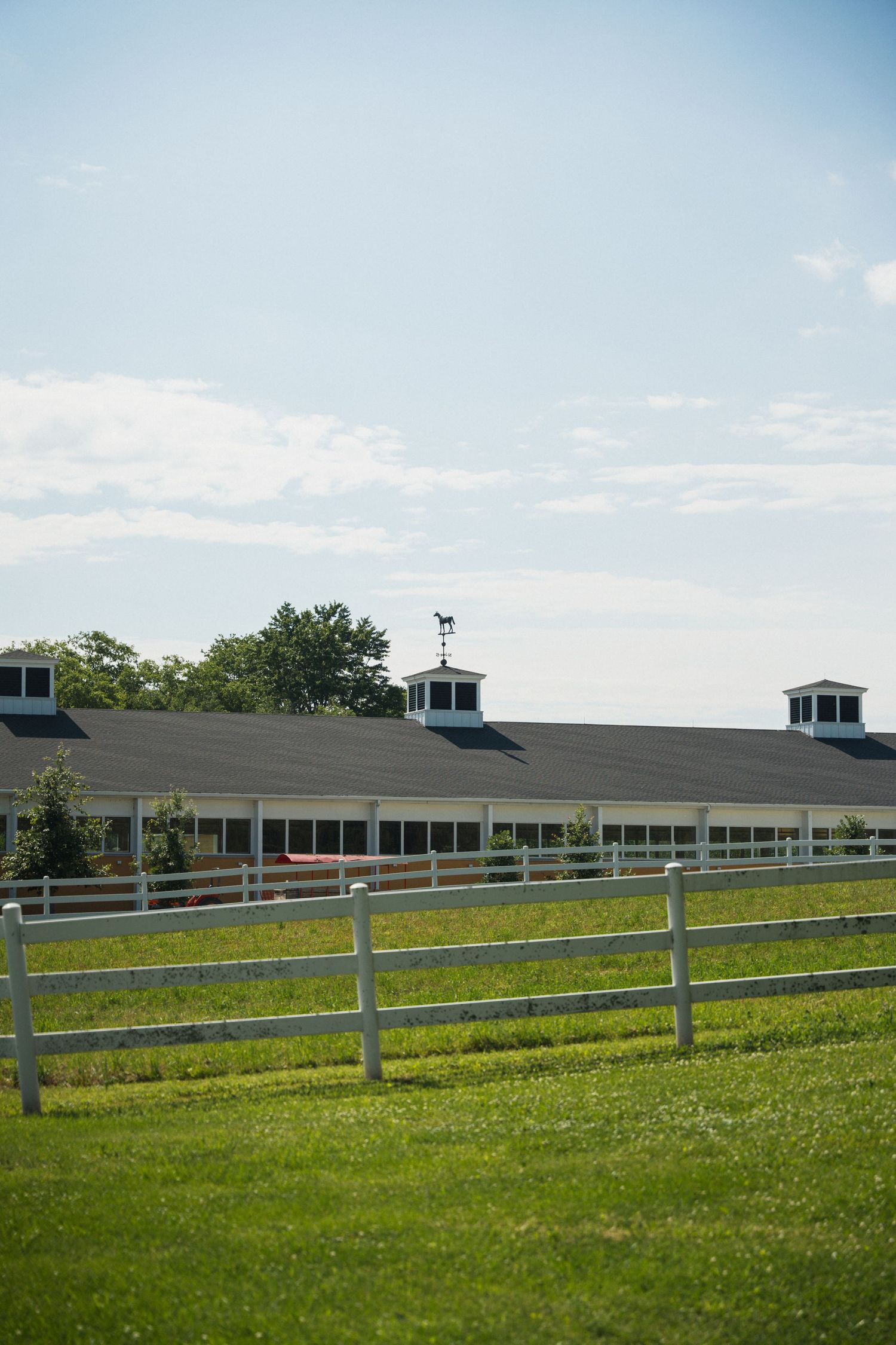 A white-fenced horse stable with cupolas sits beside lush green pastures under a partly cloudy sky.