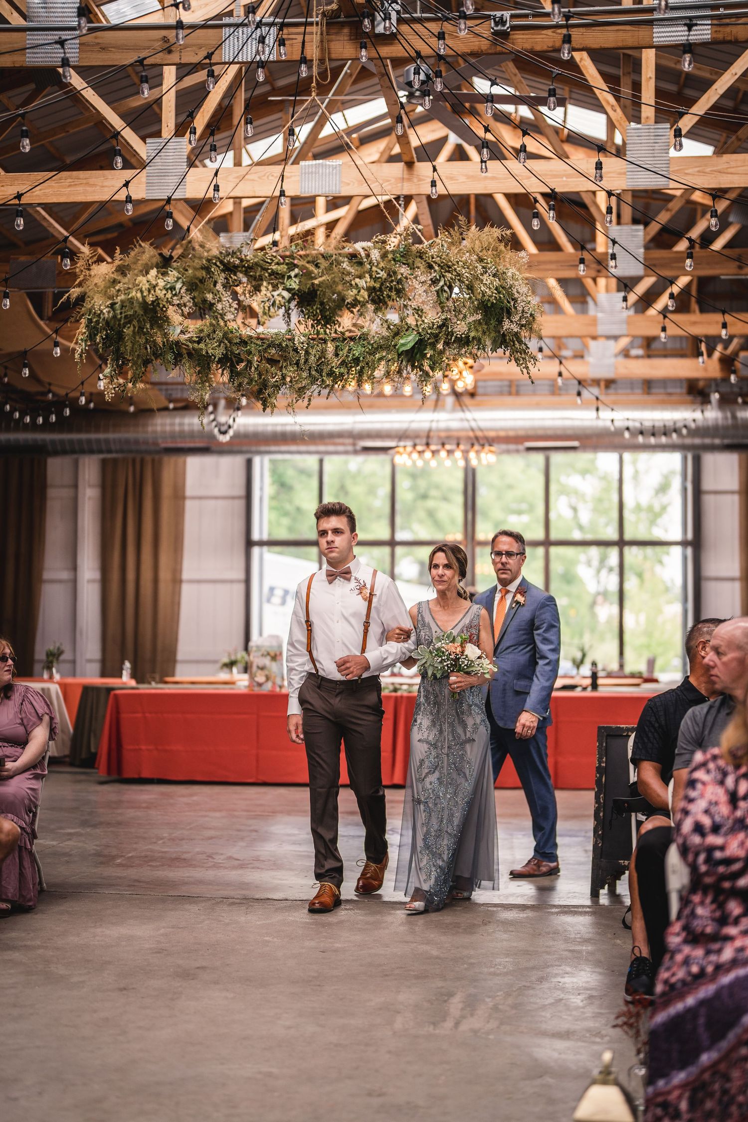 Industrial venue with hanging greenery and string lights as couple walks down aisle.