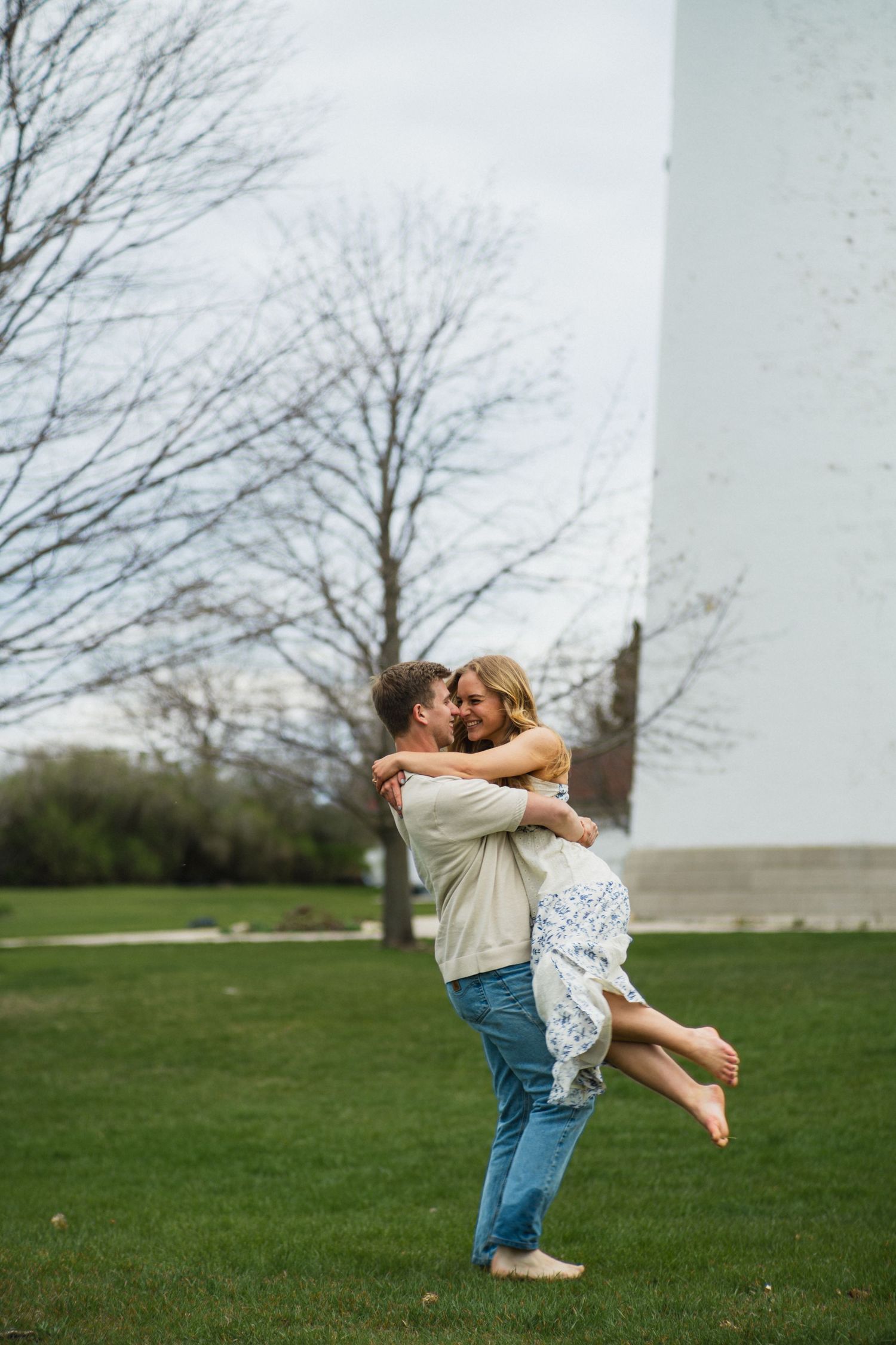 A man lifts a woman in his arms while they kiss near a white lighthouse on green grass.