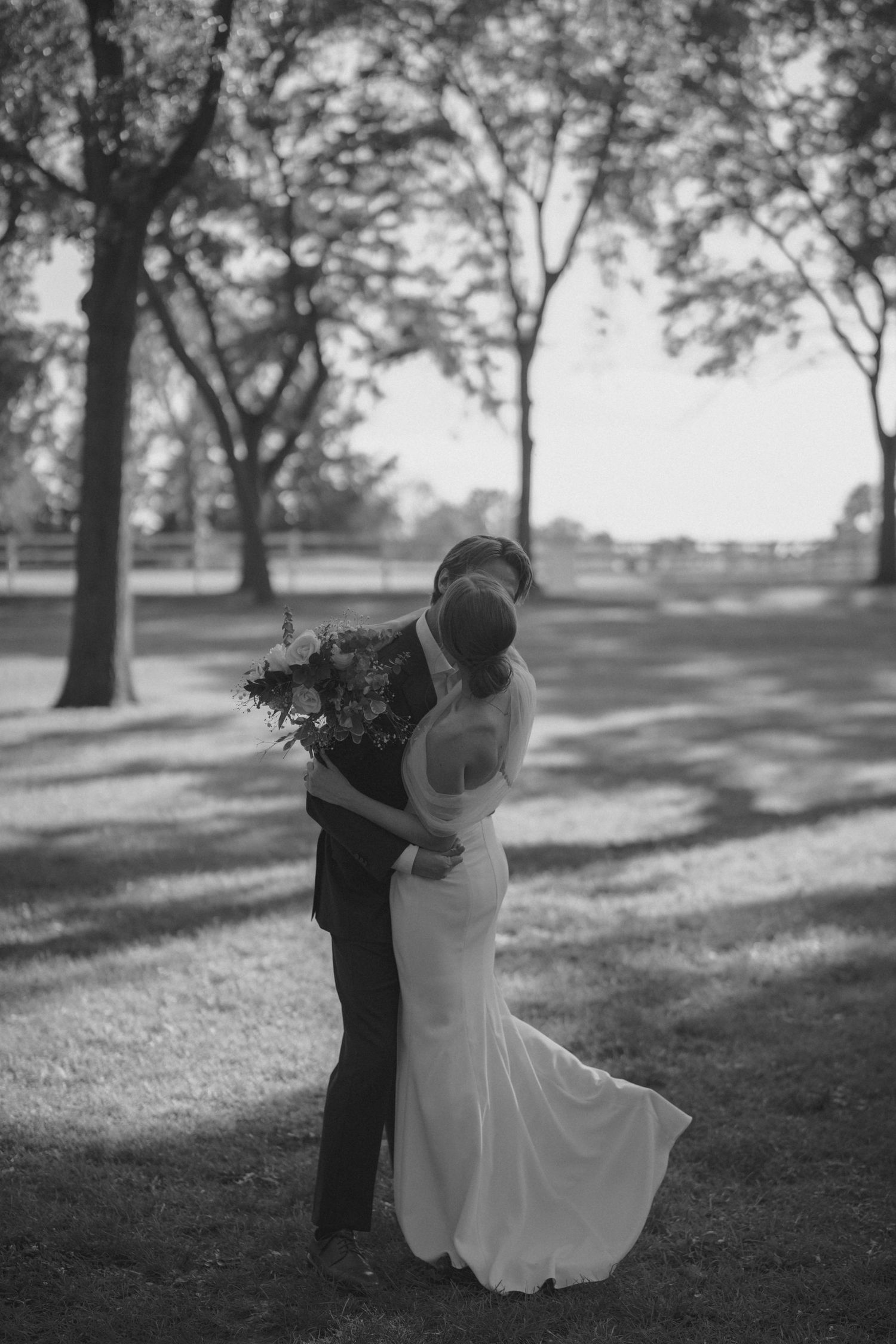 Bride and groom share romantic kiss in black and white photo surrounded by trees at outdoor wedding.