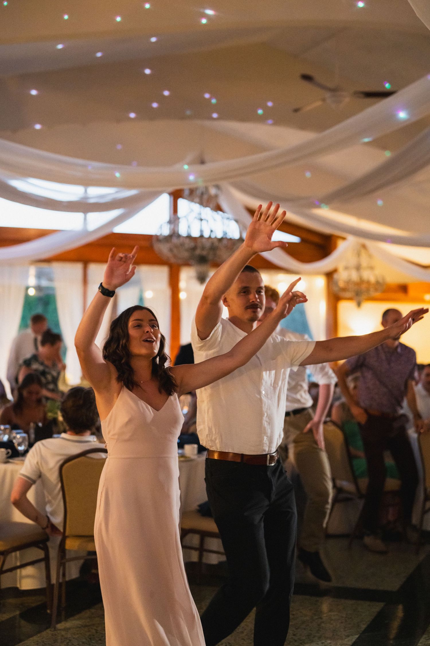 Bride and groom dance energetically with arms raised at festive wedding reception in decorated venue.