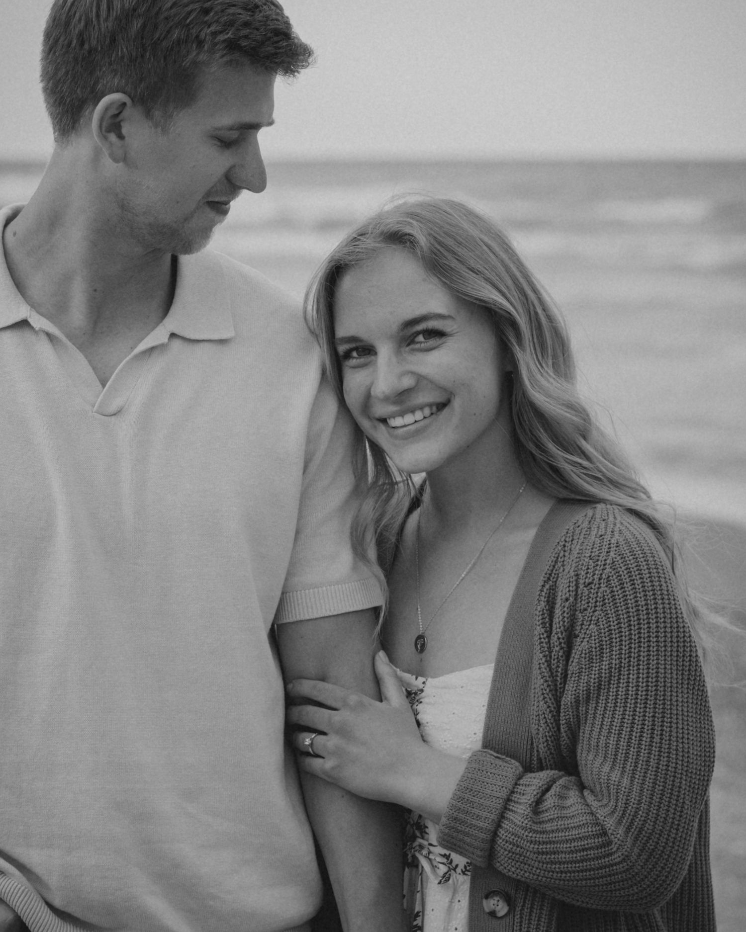 Black and white photo of a smiling couple embracing on a beach, woman in cardigan looks at camera.