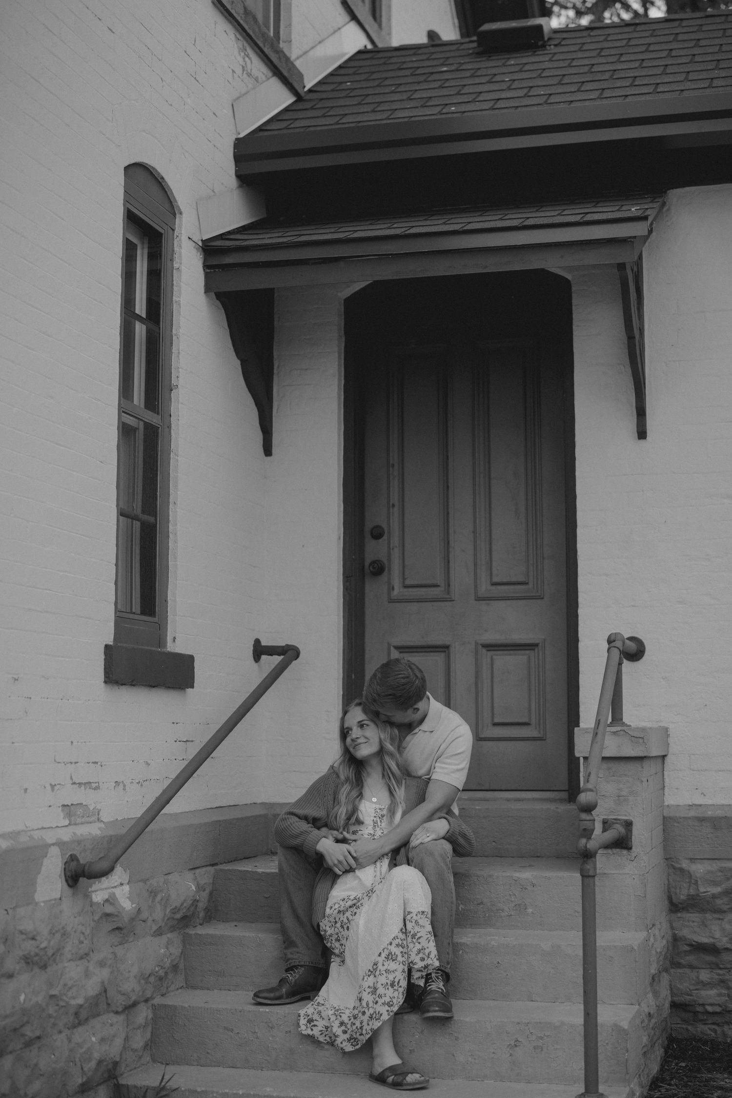 Black and white photo of couple sitting closely together on front steps of historic brick building.