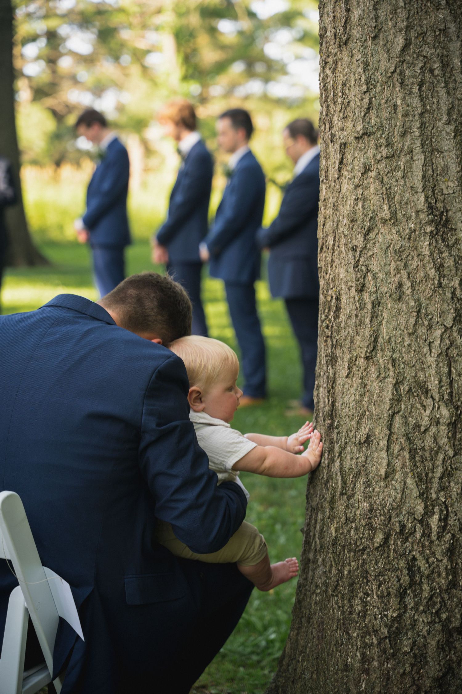 Man in navy suit holds baby touching tree bark at outdoor wedding ceremony with groomsmen in background.