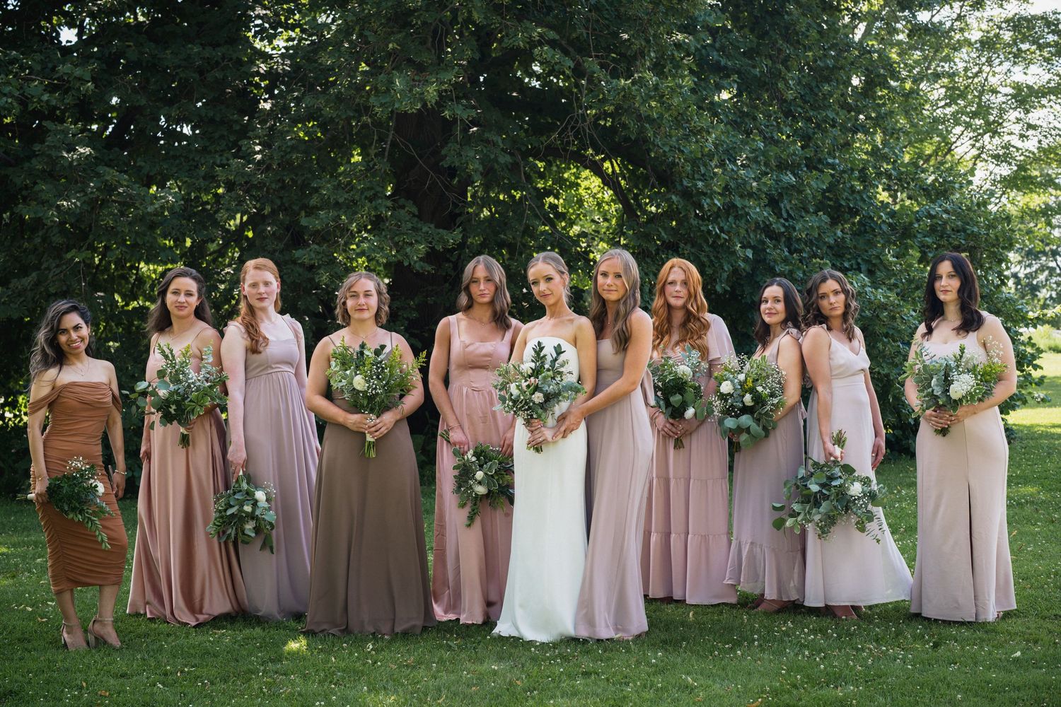 Bride poses with nine bridesmaids in mismatched pink and mauve dresses holding greenery bouquets.