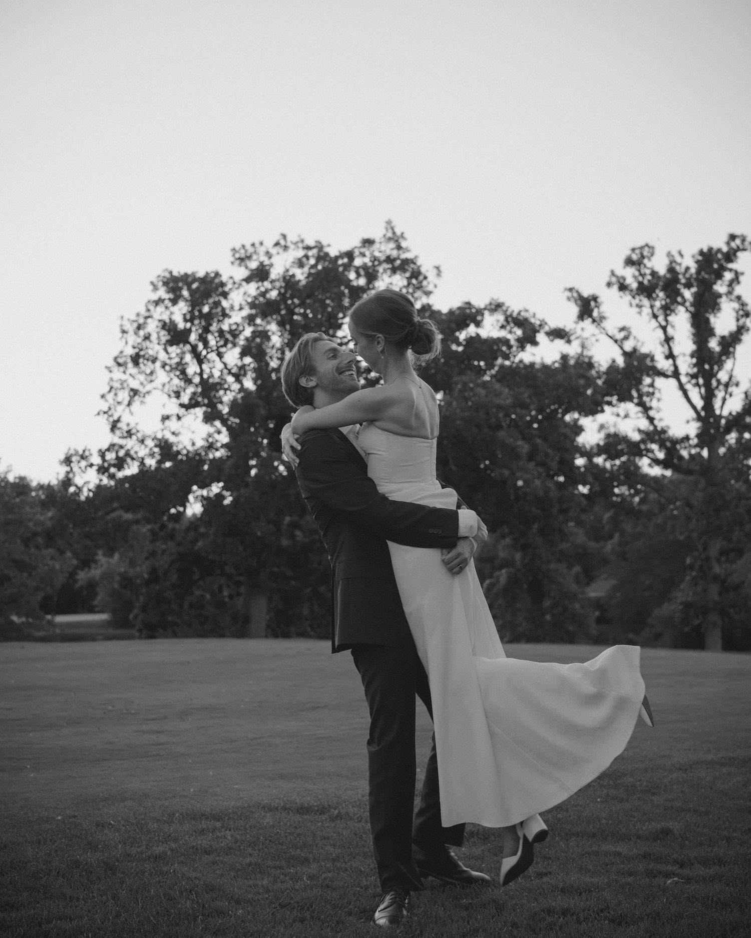 Black and white photo of groom lifting bride at sunset on lawn.
