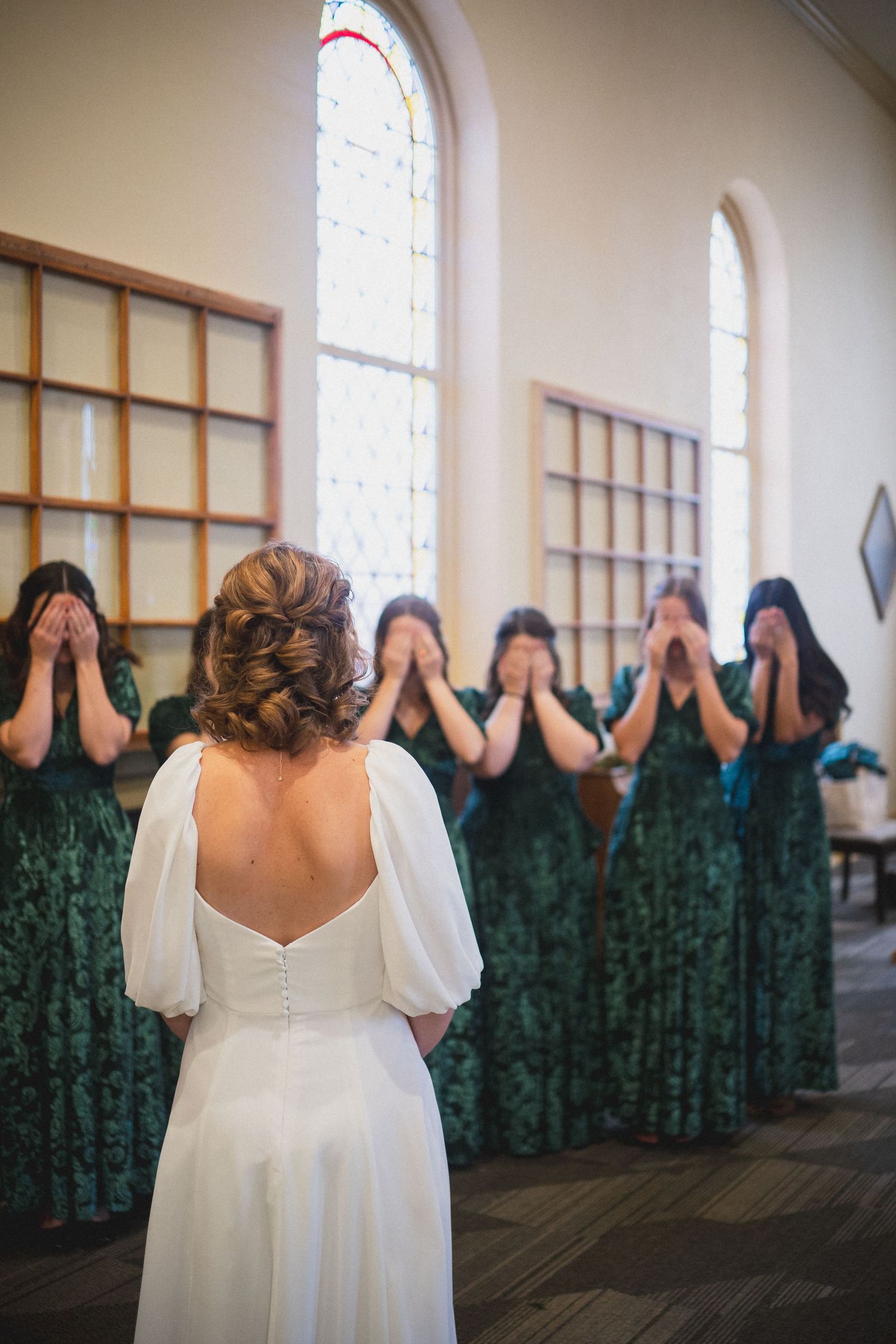 Wedding party in emerald green dresses and white gown pose playfully covering their faces in a church setting.