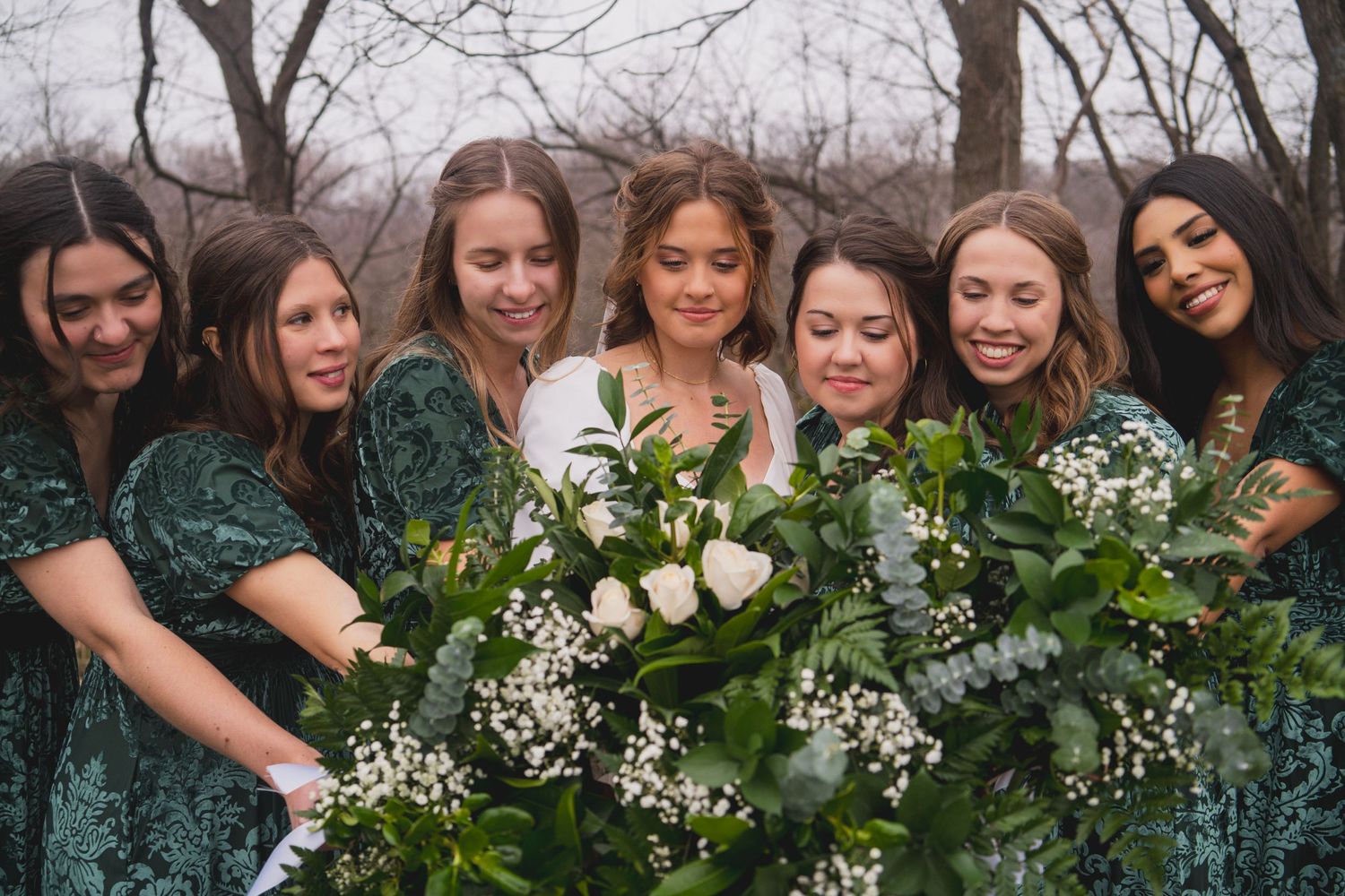 Bridesmaids gather close around bride showing off their collective bouquets of white flowers and greenery.