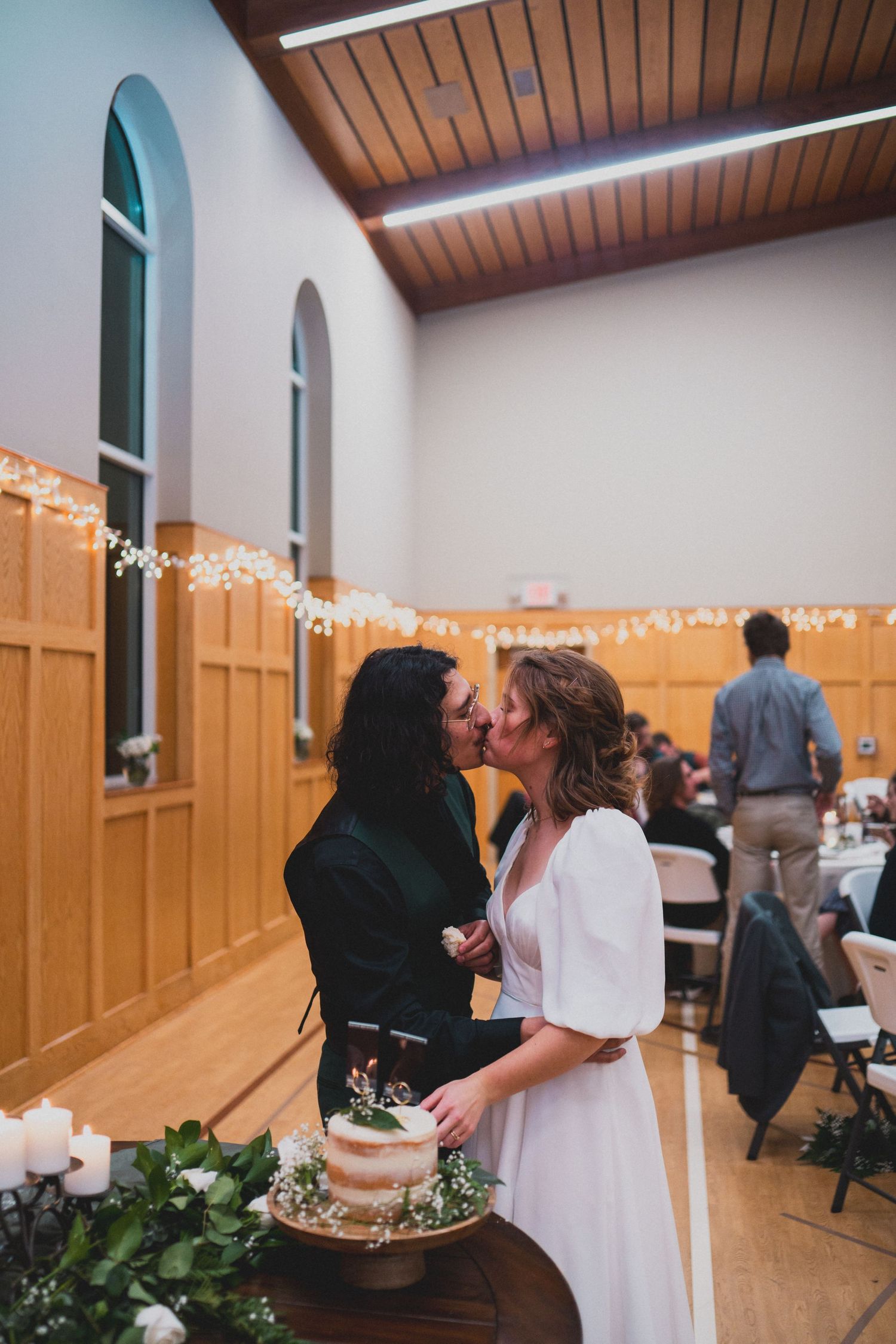 A couple shares a tender moment by their wedding cake in a reception hall with string lights.