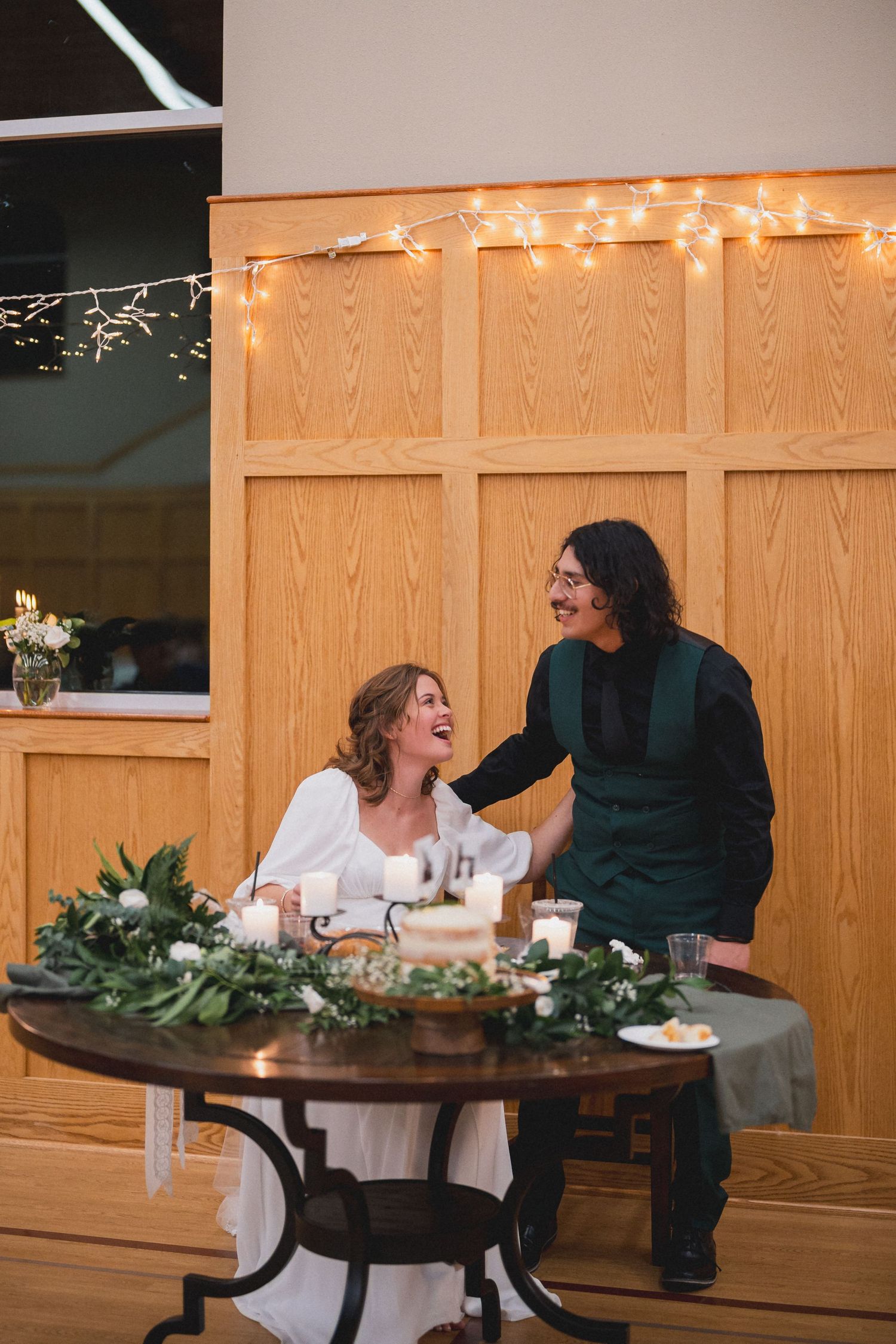 Two people share a joyful moment at a decorated table with greenery and string lights.
