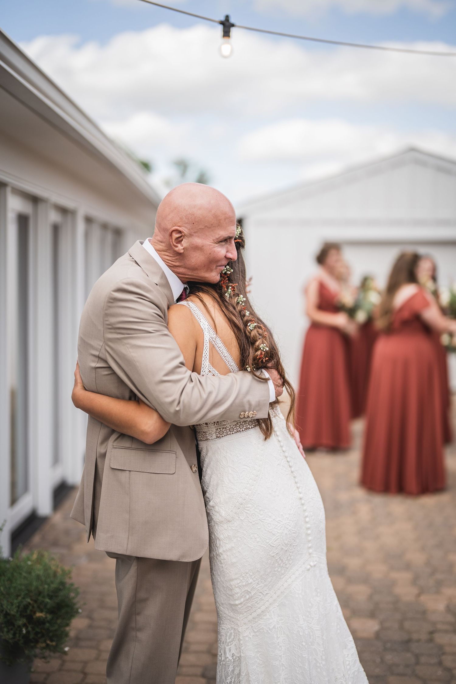 Tender embrace during outdoor wedding with bridesmaids in background wearing rust-colored dresses.