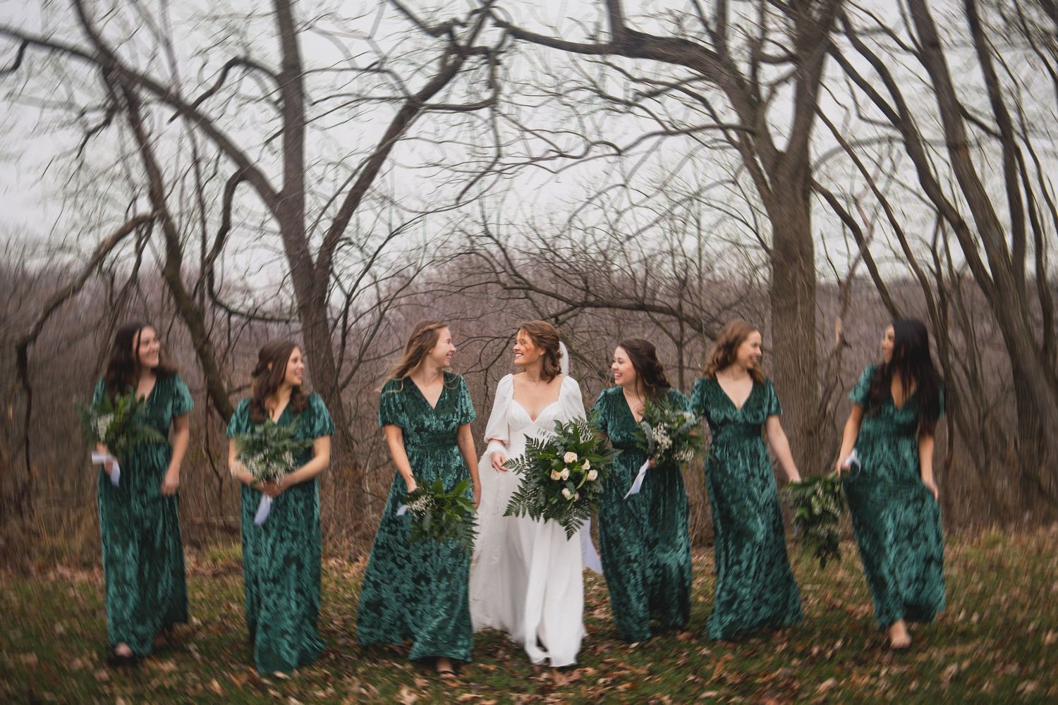 Bridal party in velvet emerald dresses stands in wooded setting with white bouquets.