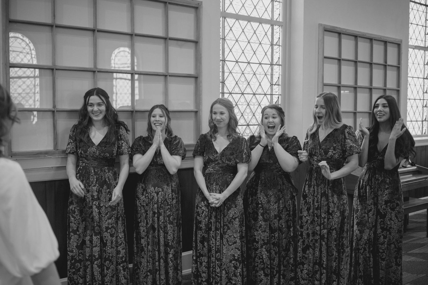 Wedding party in emerald dresses stands in line against church windows with hands covering faces.