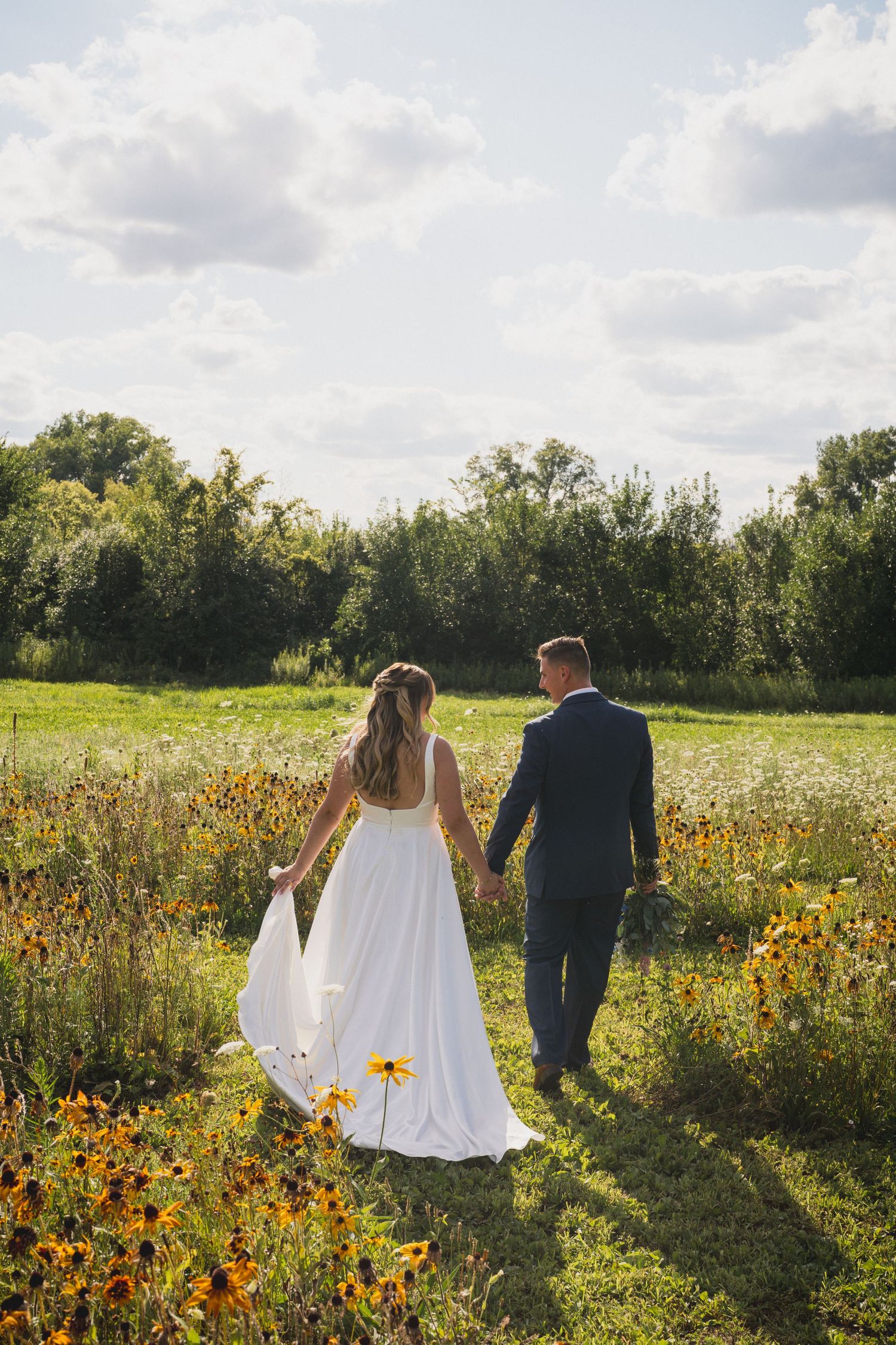 Couple walking through wildflower meadow in wedding attire at sunset.
