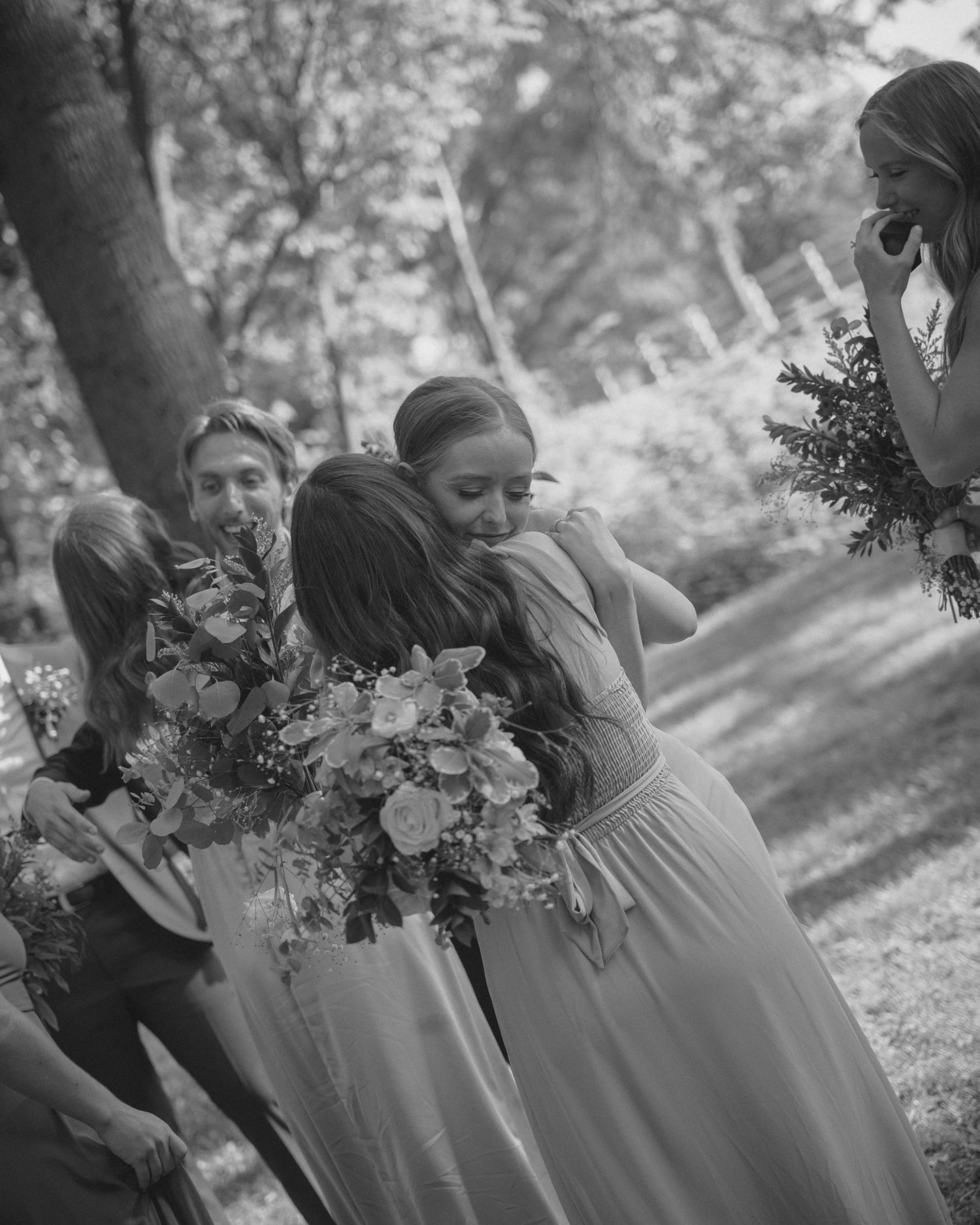 Black and white photo of bridesmaids in flowing gowns embracing and holding floral bouquets outdoors at a wedding.