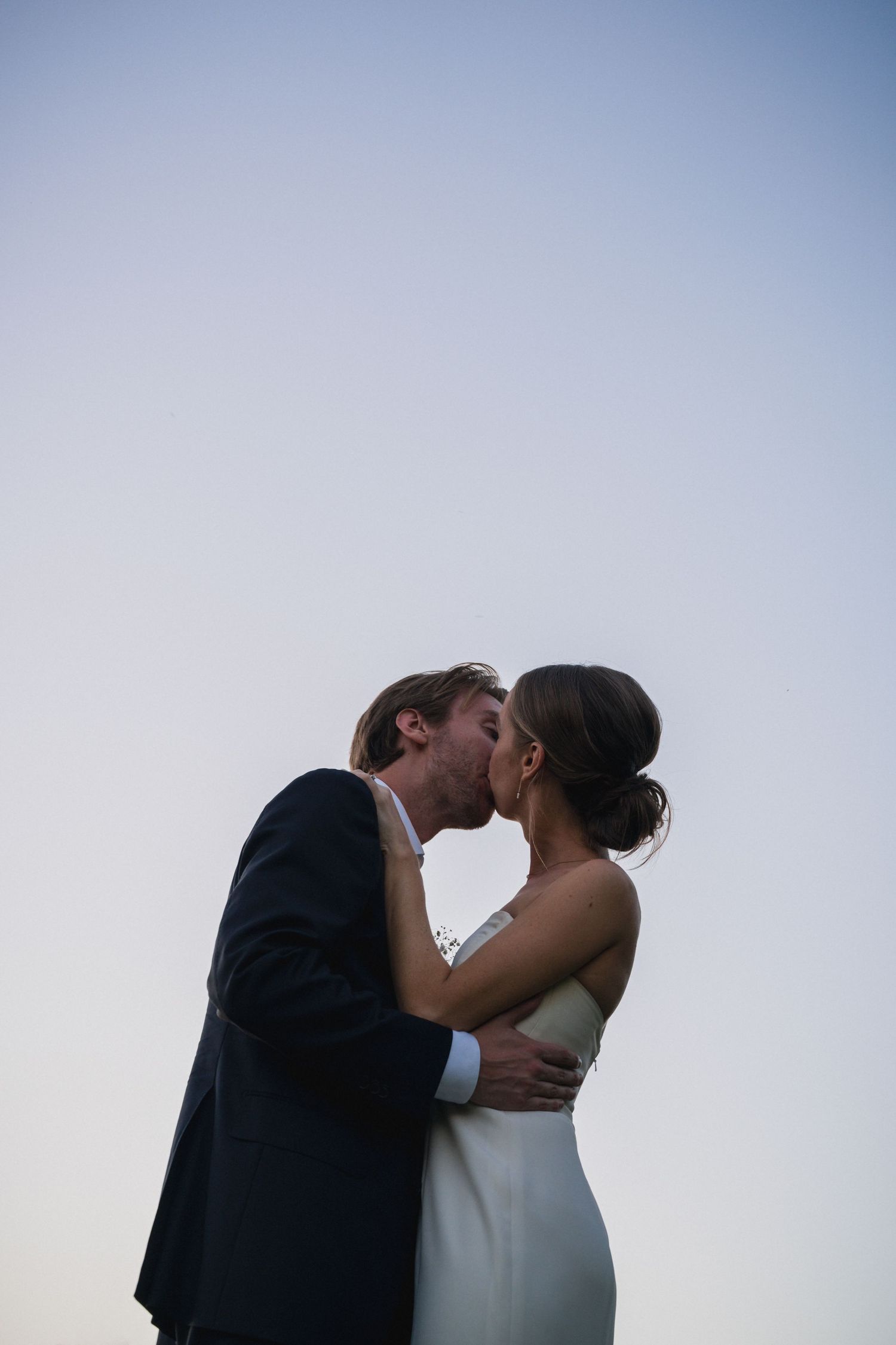 Bride and groom share a romantic kiss against a soft blue sky at their outdoor wedding ceremony.