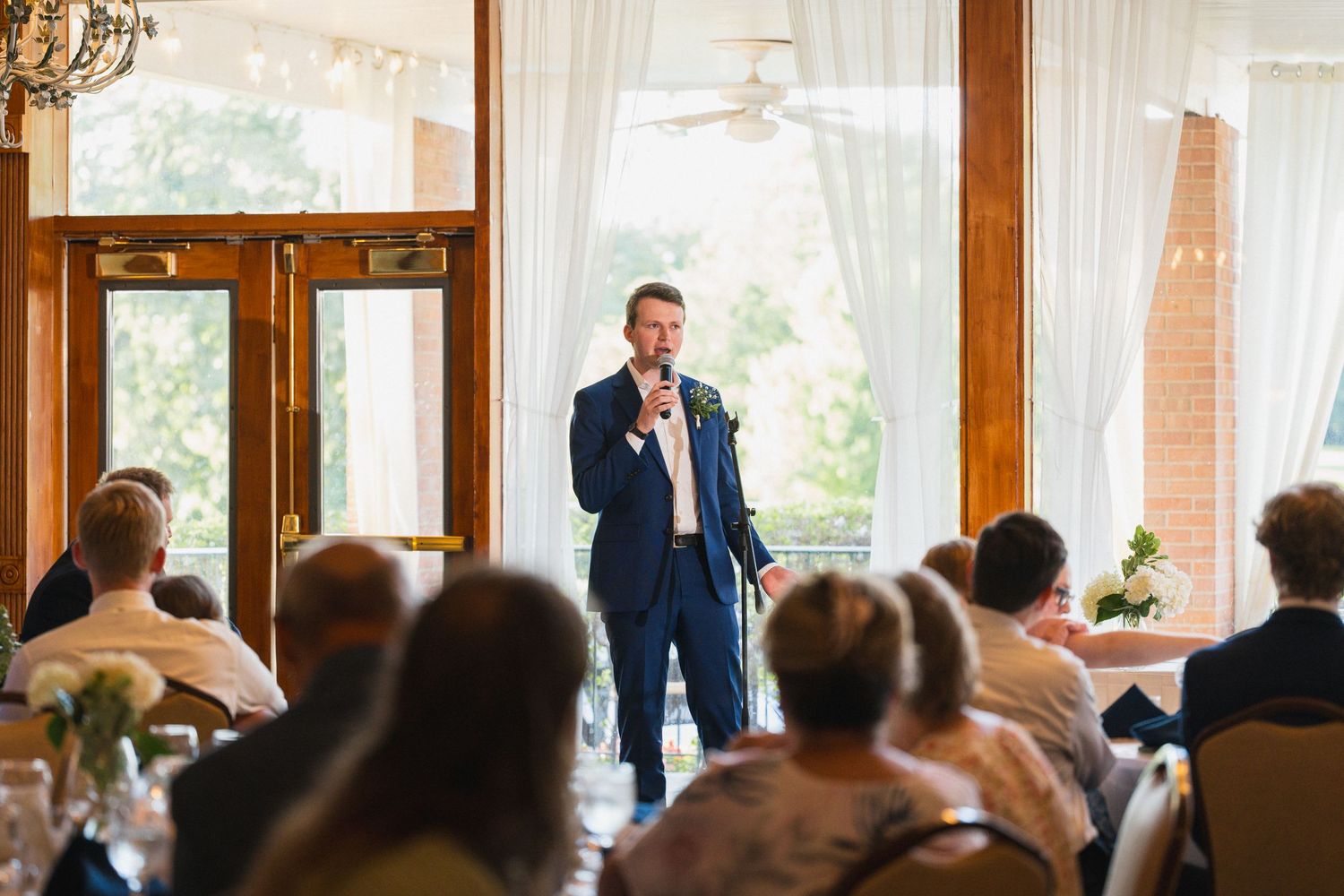 A man in a navy suit delivers a heartfelt speech at a wedding reception to seated guests.