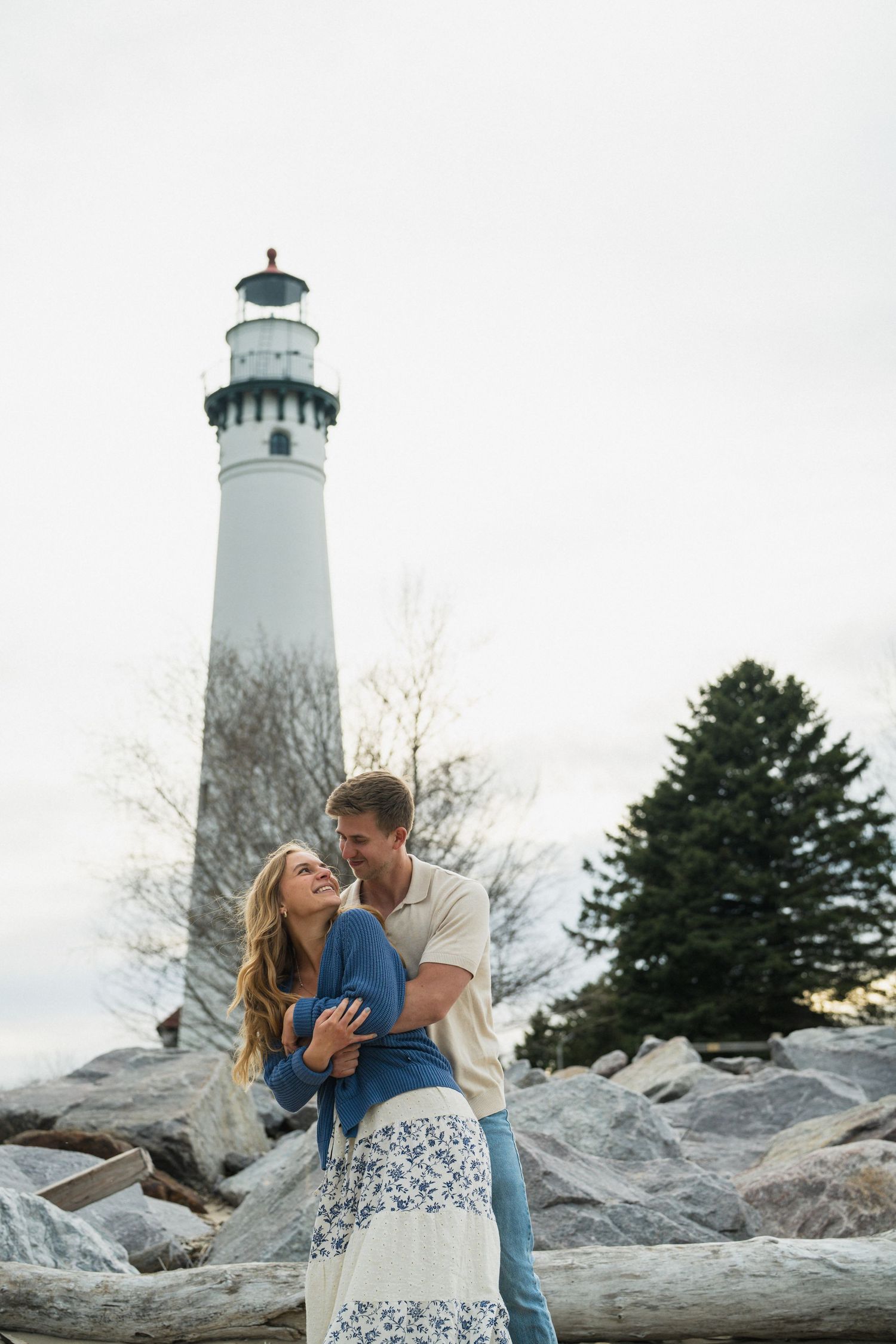 Couple embracing romantically in front of a tall white lighthouse on a rocky shore.