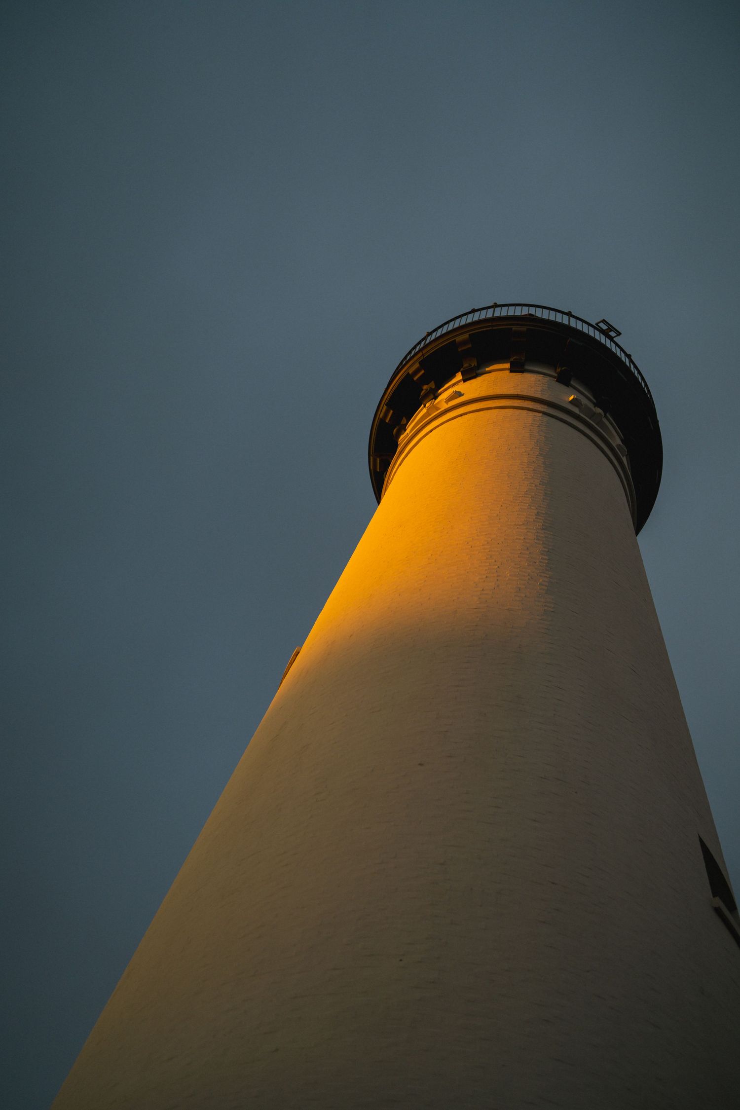 Low angle view of white lighthouse tower illuminated by warm golden light against dark sky.