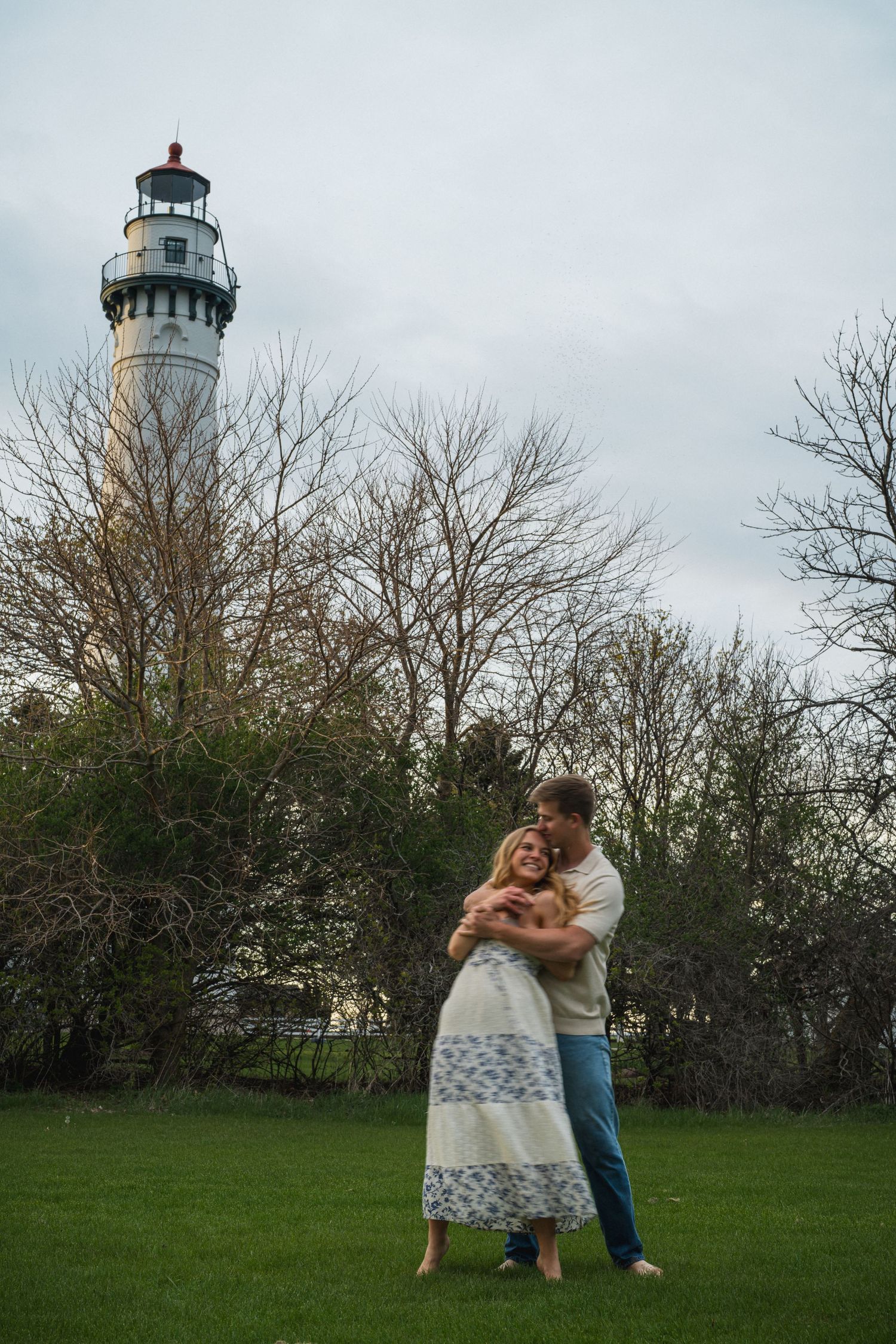Couple embracing barefoot on green grass with tall white lighthouse and bare trees in background.