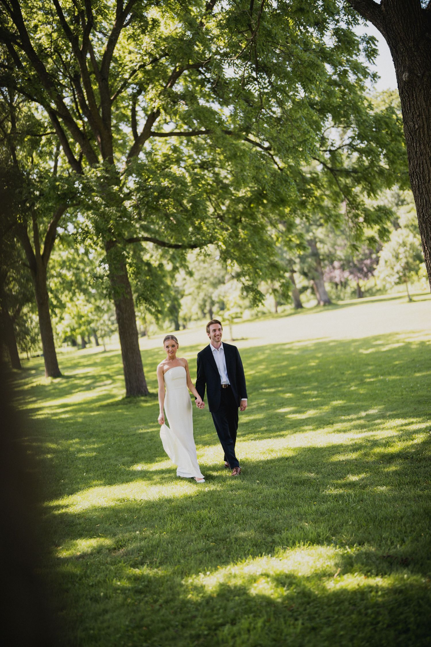 Bride and groom walk hand in hand across lush green park lawn.