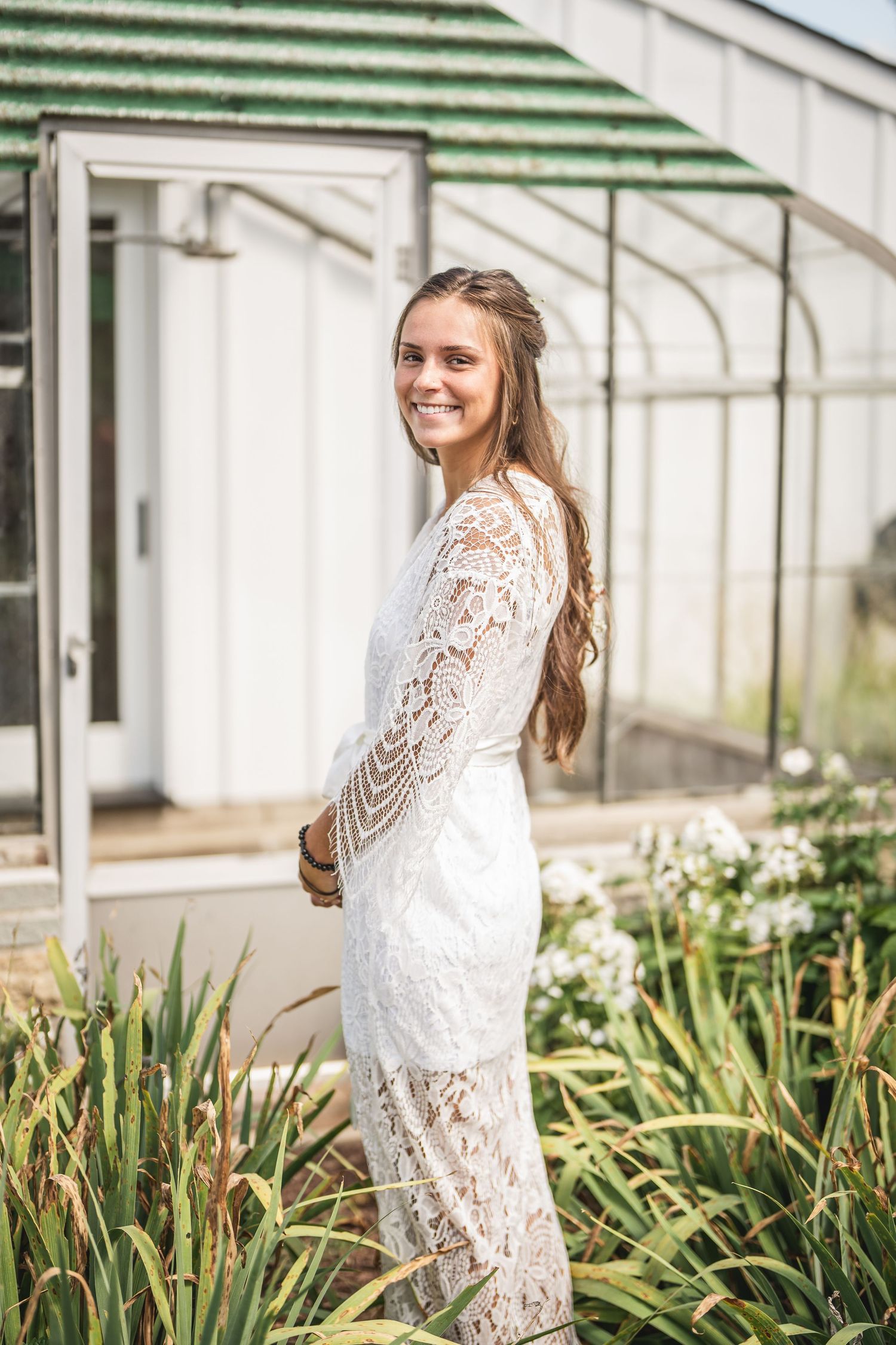Woman in white lace dress poses in front of a greenhouse with plants.