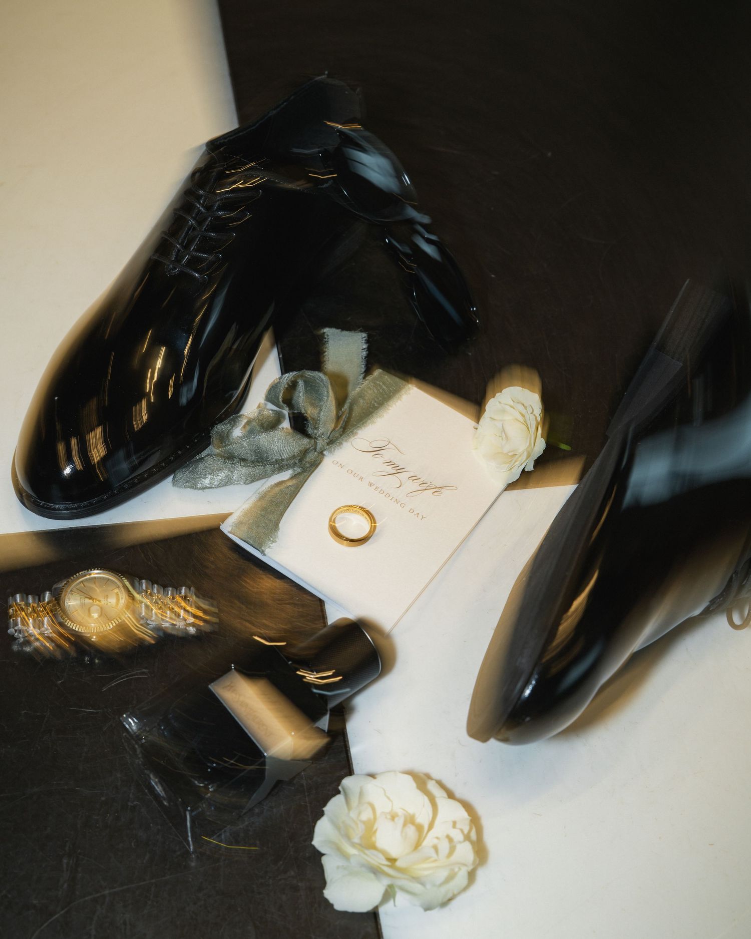 Artistic still life of black patent shoes with wedding invitation and white rose.