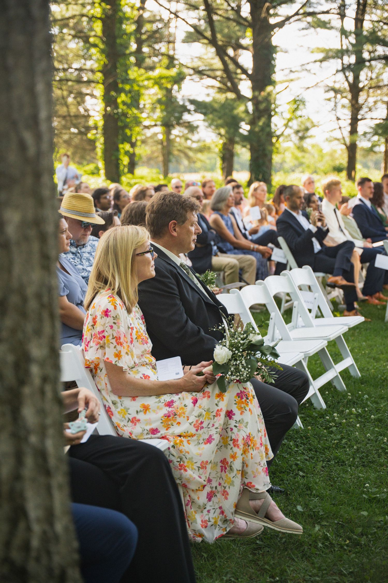 Wedding guests seated in white chairs at outdoor ceremony, woman in floral dress holding flowers.