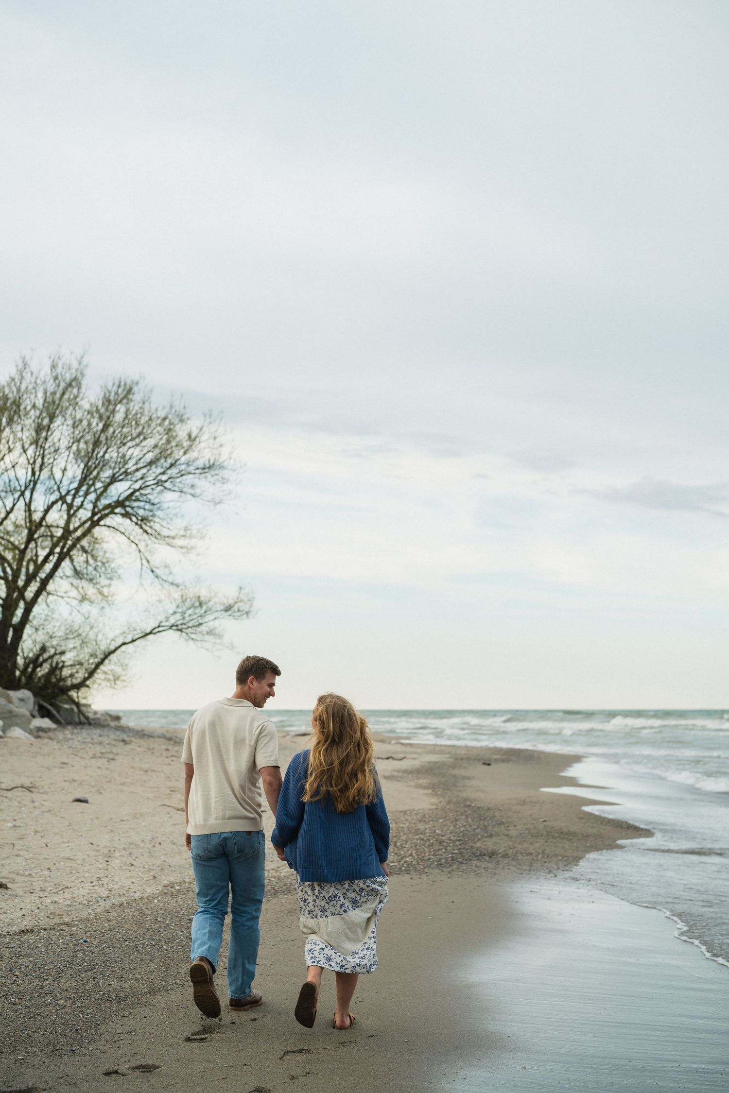 Couple walking hand in hand along a sandy beach shoreline with gentle waves and bare trees on a cloudy day.