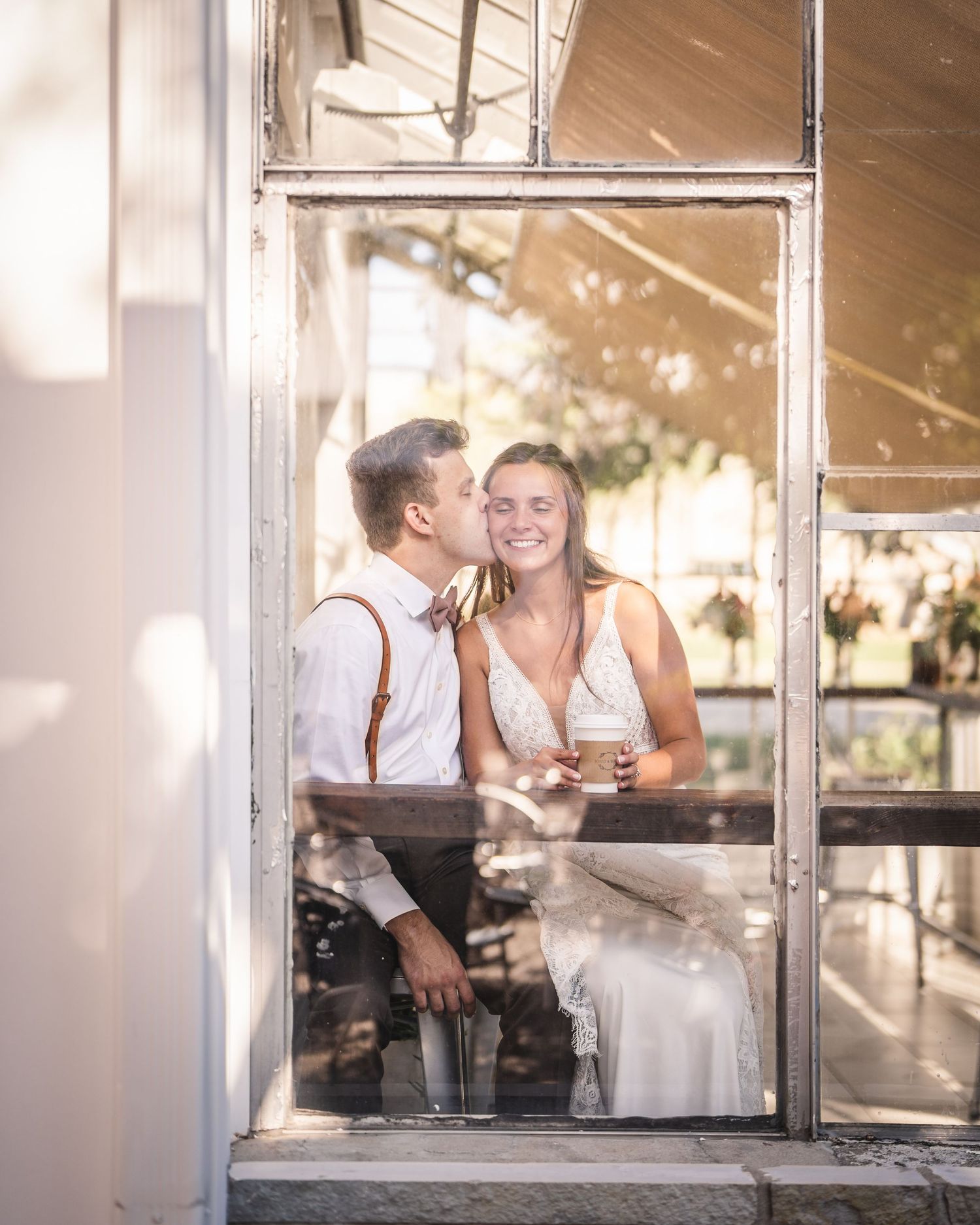 Wedding couple shares intimate moments through a vintage window frame in warm sunlight.