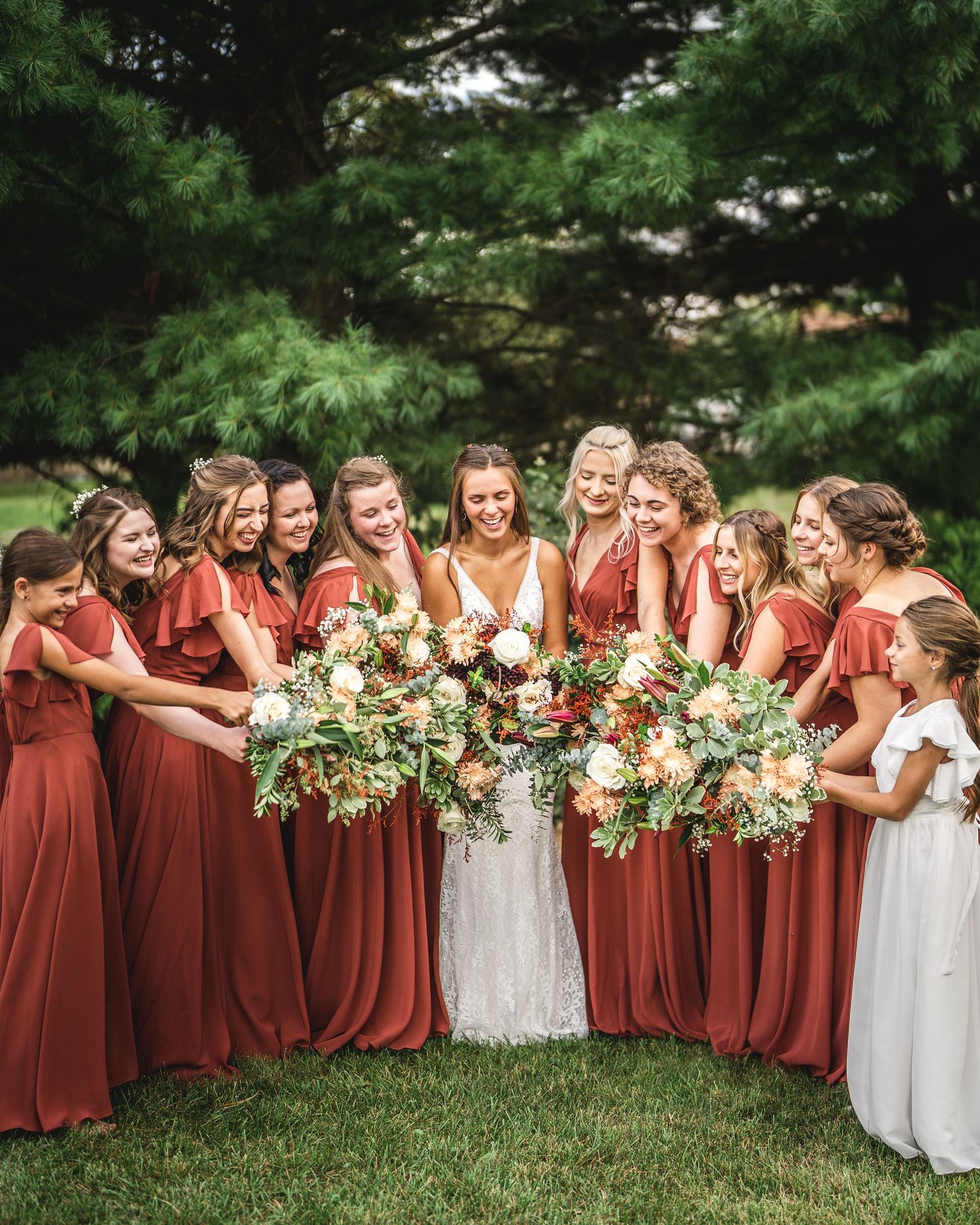 Joyful wedding party with bridesmaids in rust dresses holding floral bouquets outdoors.