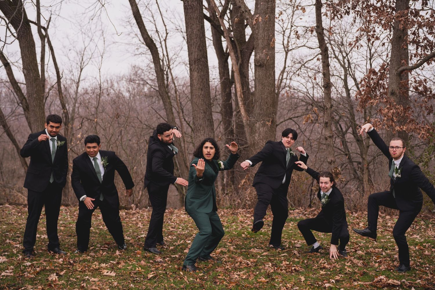 Groomsmen strike fun poses in black suits with one member in emerald green outdoors.