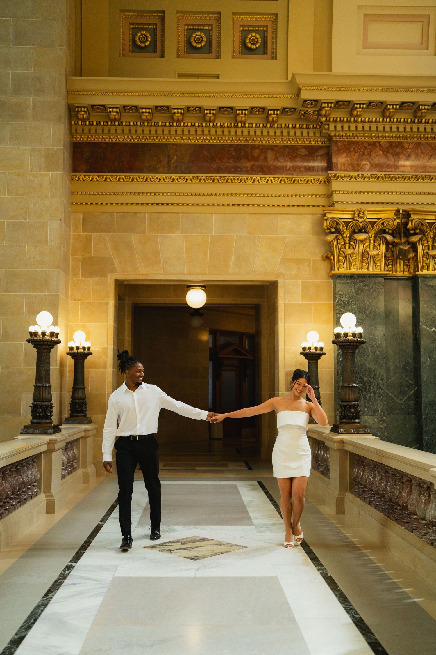 Couple in formal attire walking through elegant marble hallway with ornate architecture.