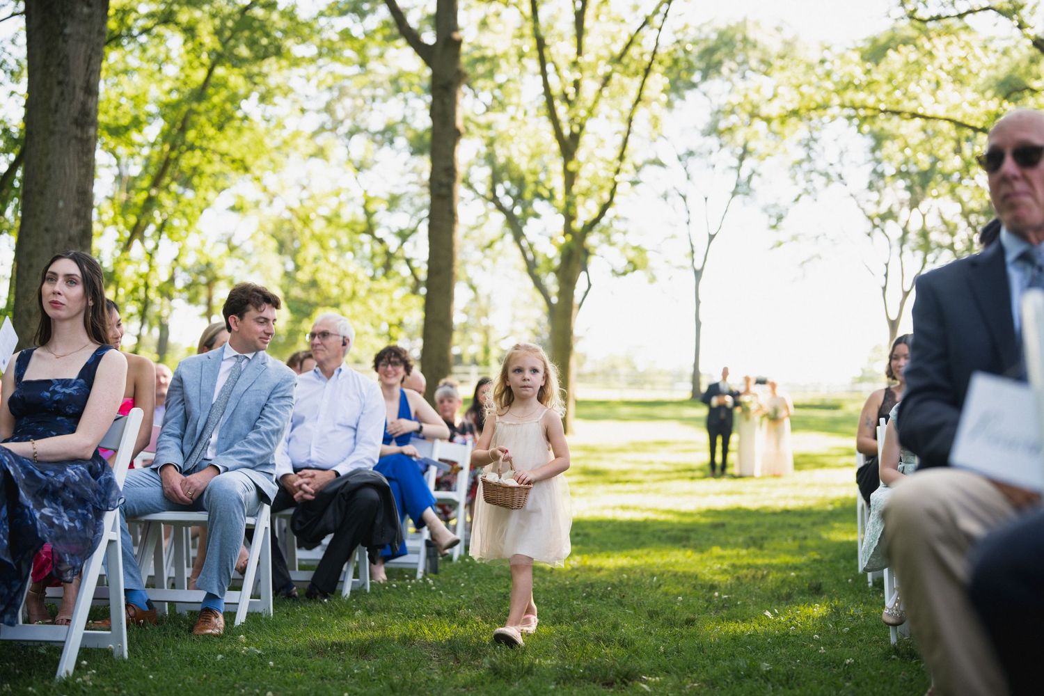 Flower girl walking down outdoor wedding ceremony aisle with seated guests watching.