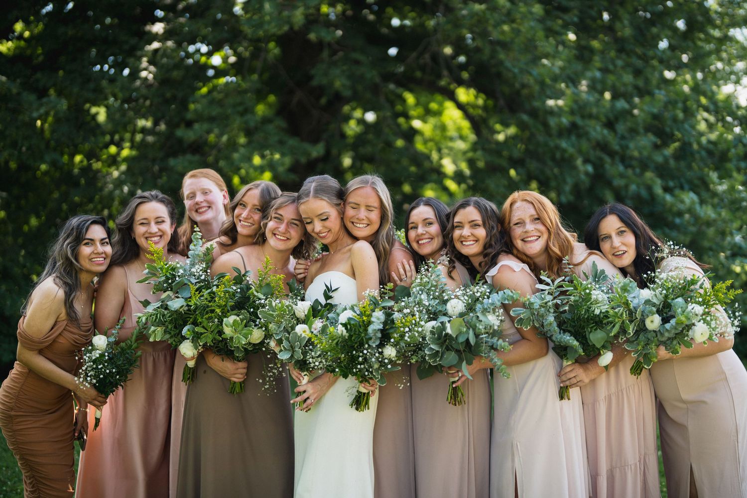 Bride and bridesmaids in mismatched neutral dresses holding white and green bouquets outdoors.