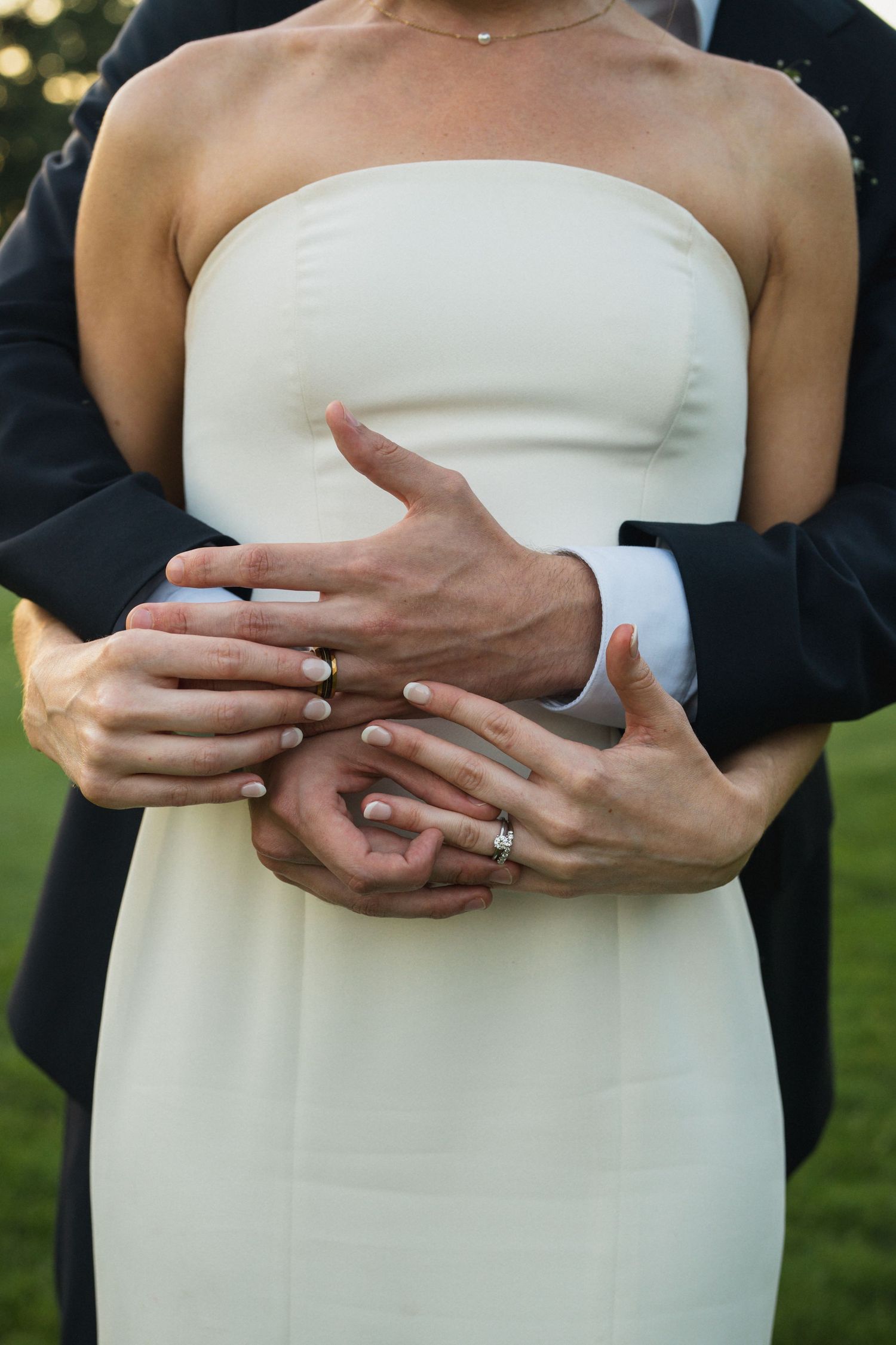 Newlyweds display wedding rings as groom embraces bride in strapless white dress from behind outdoors.
