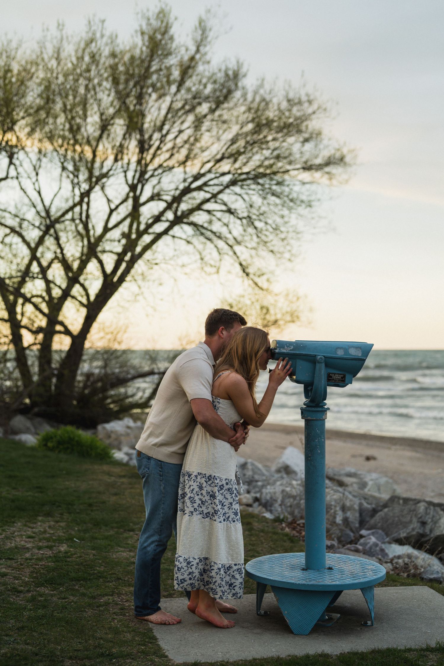 Couple sharing romantic kiss beside vintage coin-operated binoculars at sunset near rocky beach shoreline.