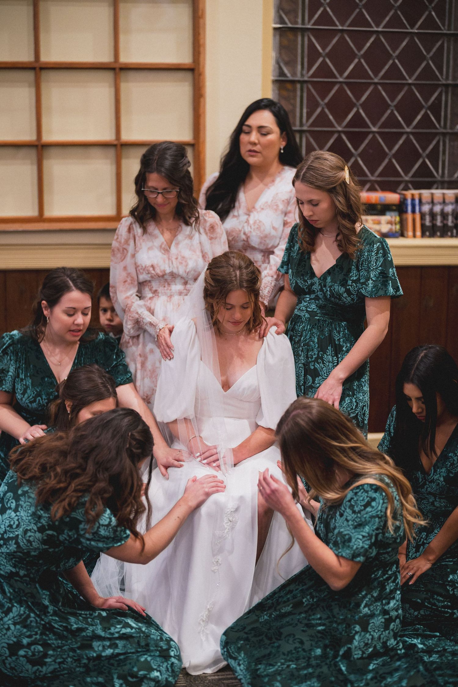 Bridesmaids in emerald green dresses gather around the bride in prayer.