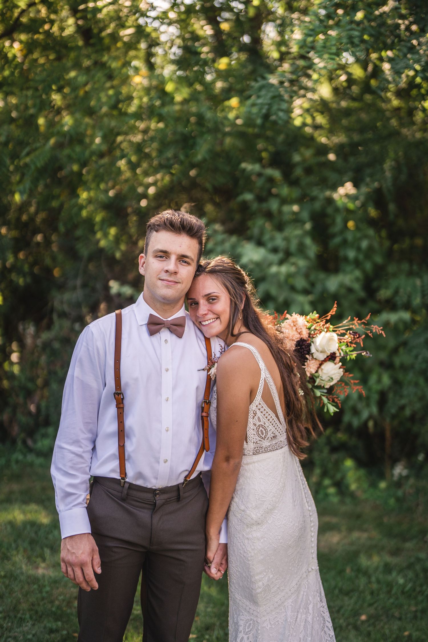 A couple poses for wedding photos in a garden setting wearing rustic-themed wedding attire.