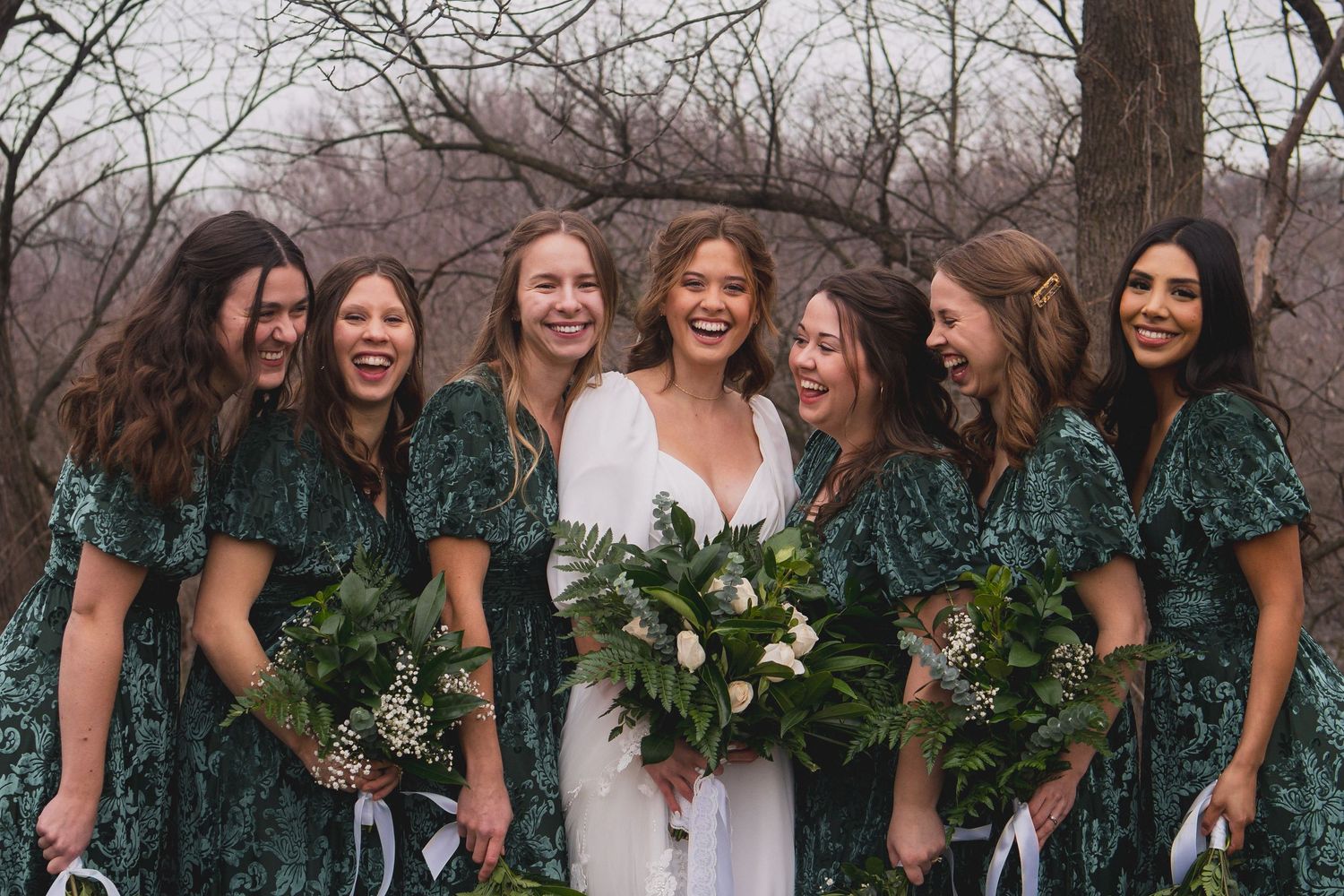 Joyful bridal party laughing together while holding lush green bouquets outdoors.